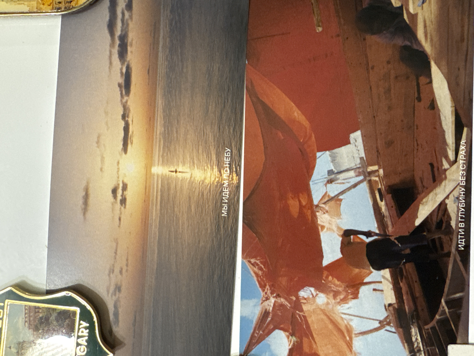 The image shows two vertically oriented postcards placed side-by-side. The postcard on the left depicts a sunset over water with a silhouette of a small boat in the distance. Text, in Russian, is visible on the right side of the card. The postcard on the right depicts the interior of a wooden structure with people. Text, in Russian, is visible on the right side of the card. A decorative metal object with the word "HUNGARY" is present on the lower left side of the image. There is no information about Kazan or Russia.