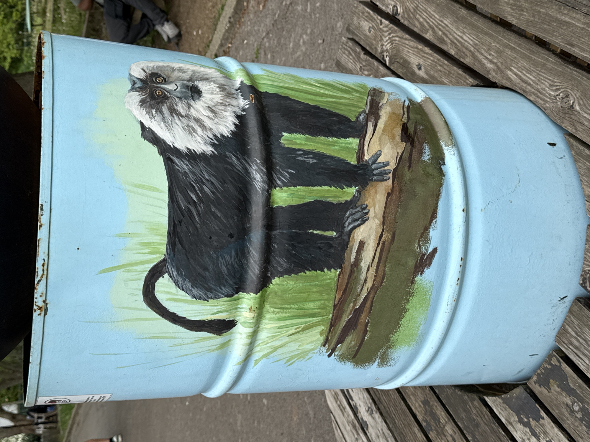 A cylindrical metal trash can, painted light blue, sits outdoors on wooden planks, possibly a bench. The can is decorated with a painting of a monkey, specifically an Emperor Tamarin, perched on a brown tree branch against a green grassy background. Part of a person wearing dark pants and light sneakers is visible in the background. The location is likely in the city of Sofia, Bulgaria, based on prior knowledge of the image source.