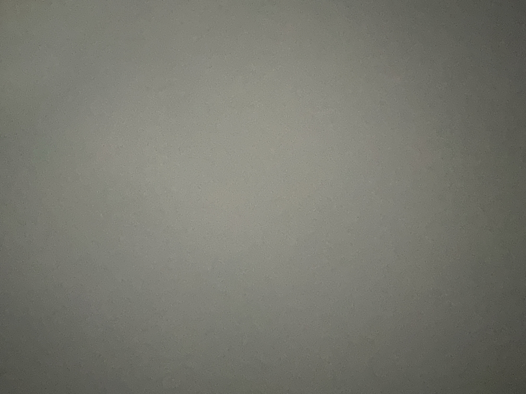 The image is a close-up, texture-heavy shot of a speckled gray surface, likely a wall or a countertop. There are no discernible subjects such as people, objects, or a specific scene. The lighting appears somewhat dim, with a gradient effect causing the top left of the image to be brighter than the bottom right, suggesting a light source from the upper left. The texture is granular, with small, dark speckles distributed evenly across the gray background. No activity or event is taking place. There are no visible location cues, time of day indicators, weather conditions, or emotions conveyed by the image. No text is present. The image is a purely abstract representation of a textured surface.