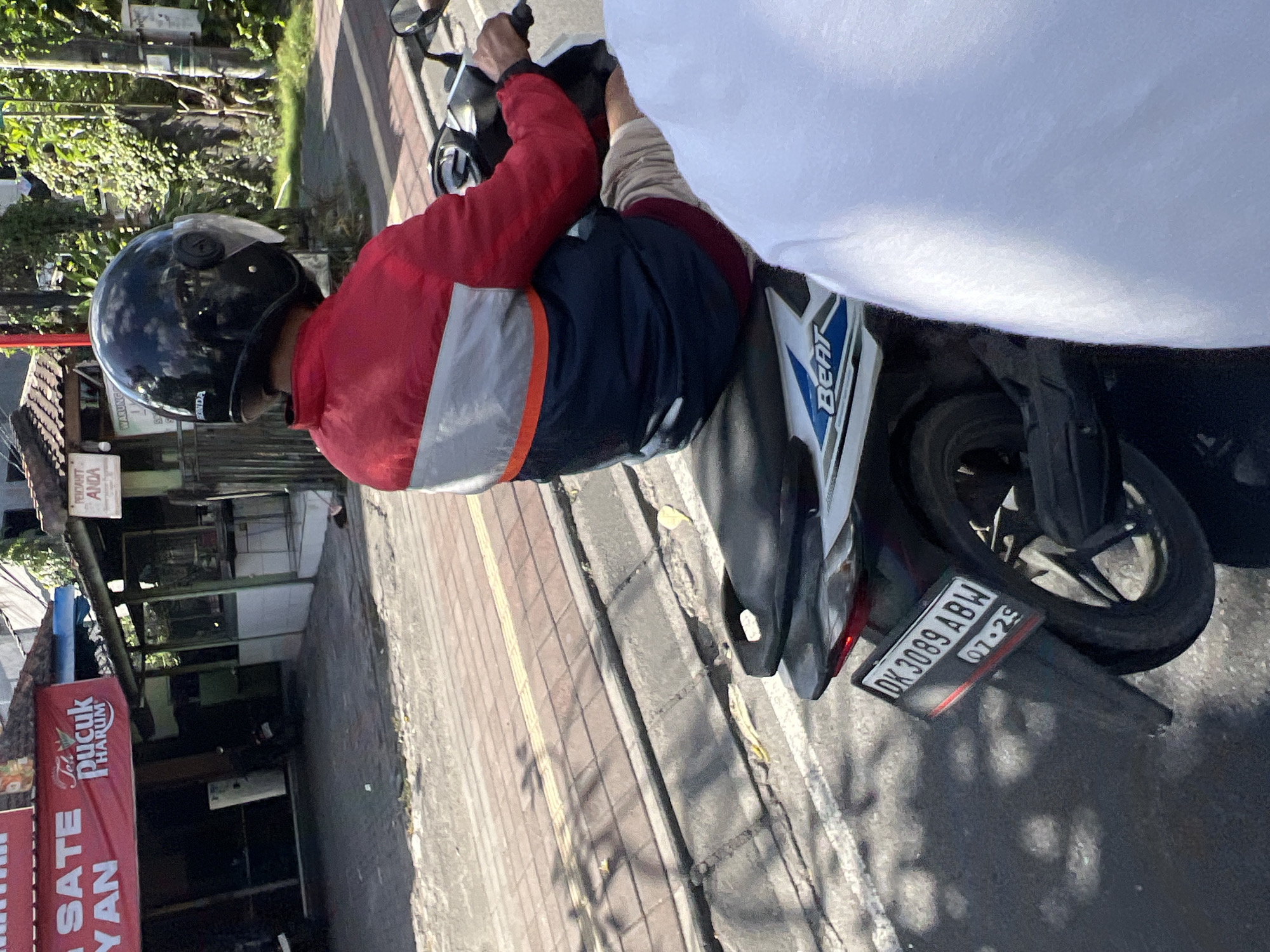A person wearing a red, white, and blue jacket and a black helmet rides a white and blue Honda Beat scooter in Kuta, Indonesia. The scooter has a license plate that reads "DK 3089 ABW" and "07.29". The rider is in motion, on a street lined with shops and trees. A red banner with white text that says "SATE Pucuk HARUM" is visible on the left side of the image. The sun is shining, casting shadows on the road.