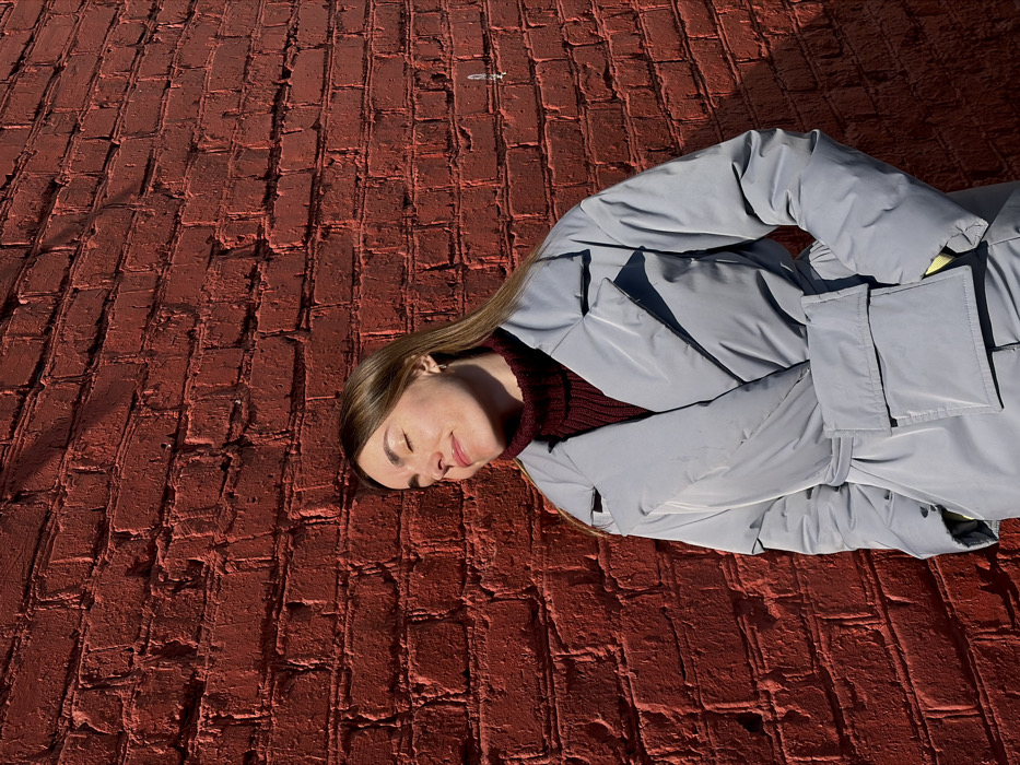 A single female subject stands facing the viewer, slightly angled towards the right side of the frame, in front of a red brick wall. Her eyes are closed, and her head is tilted upward and slightly to her right, exposing her face to direct sunlight, indicating an interaction with the sun's warmth. She has light brown hair, visible past her shoulders. A small, silver-colored hoop earring is present in her visible left ear. She is wearing a burgundy, ribbed turtleneck sweater under a light blue or pale grey shiny puffer coat. The coat features a wide collar and lapels, is cinched at the waist by a matching belt, and has a prominent rectangular flap pocket on the left side of the torso. Her hands are in the coat pockets, with a hint of a yellow garment or lining visible near her right wrist. The background consists entirely of a textured red brick wall, illuminated by strong sunlight from the upper left, casting distinct shadows on the wall and behind the subject. No specific landmarks or unique architectural features are present to precisely identify the location within Izhevsk, Russia. The scene suggests an outdoor setting on a clear, sunny day.