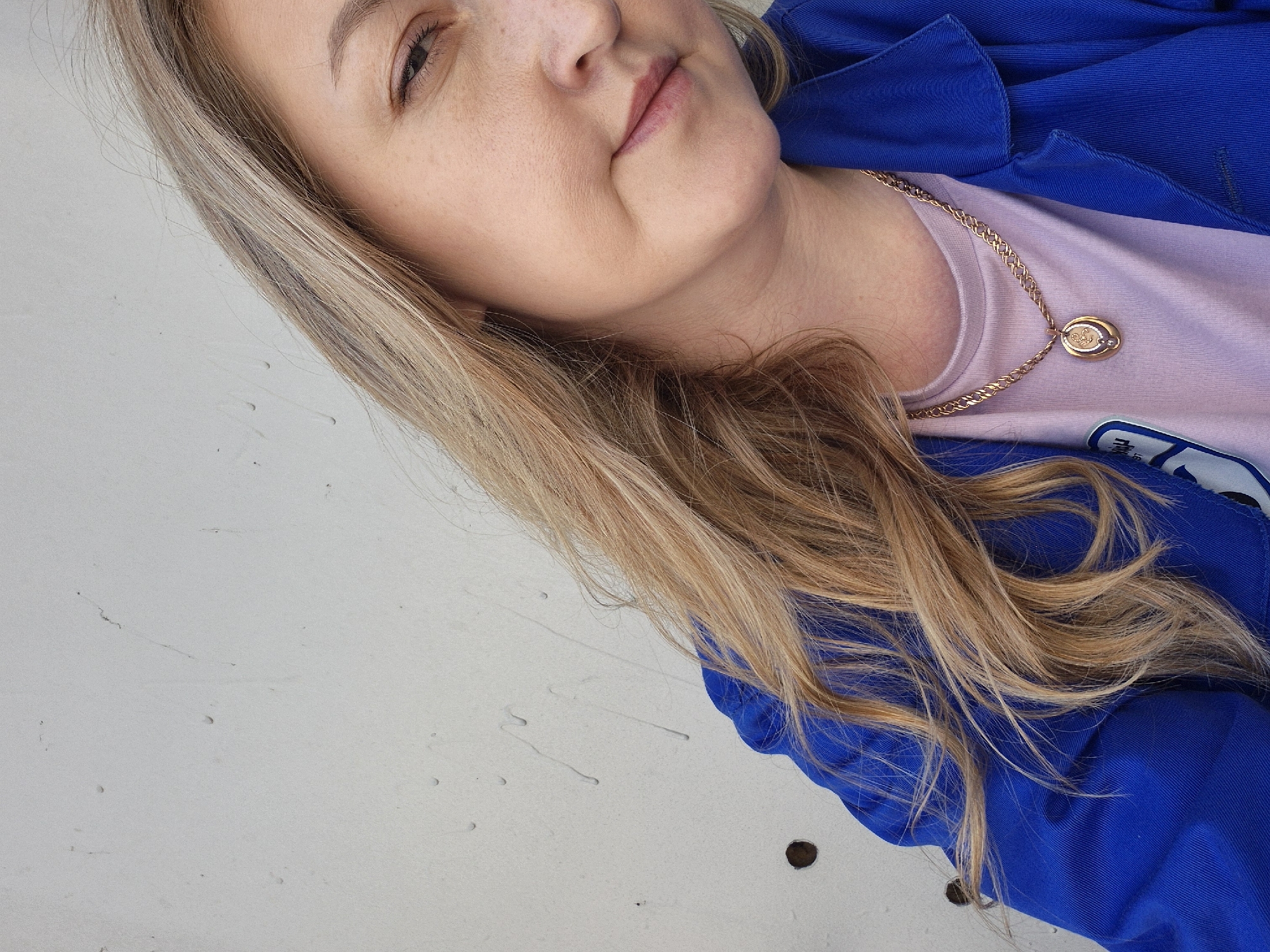 A woman with blonde hair is wearing a blue jacket and a light purple t-shirt with a gold necklace. The background appears to be a white or light-colored surface. The photo was taken in the city of Chaikovsky, Russia.