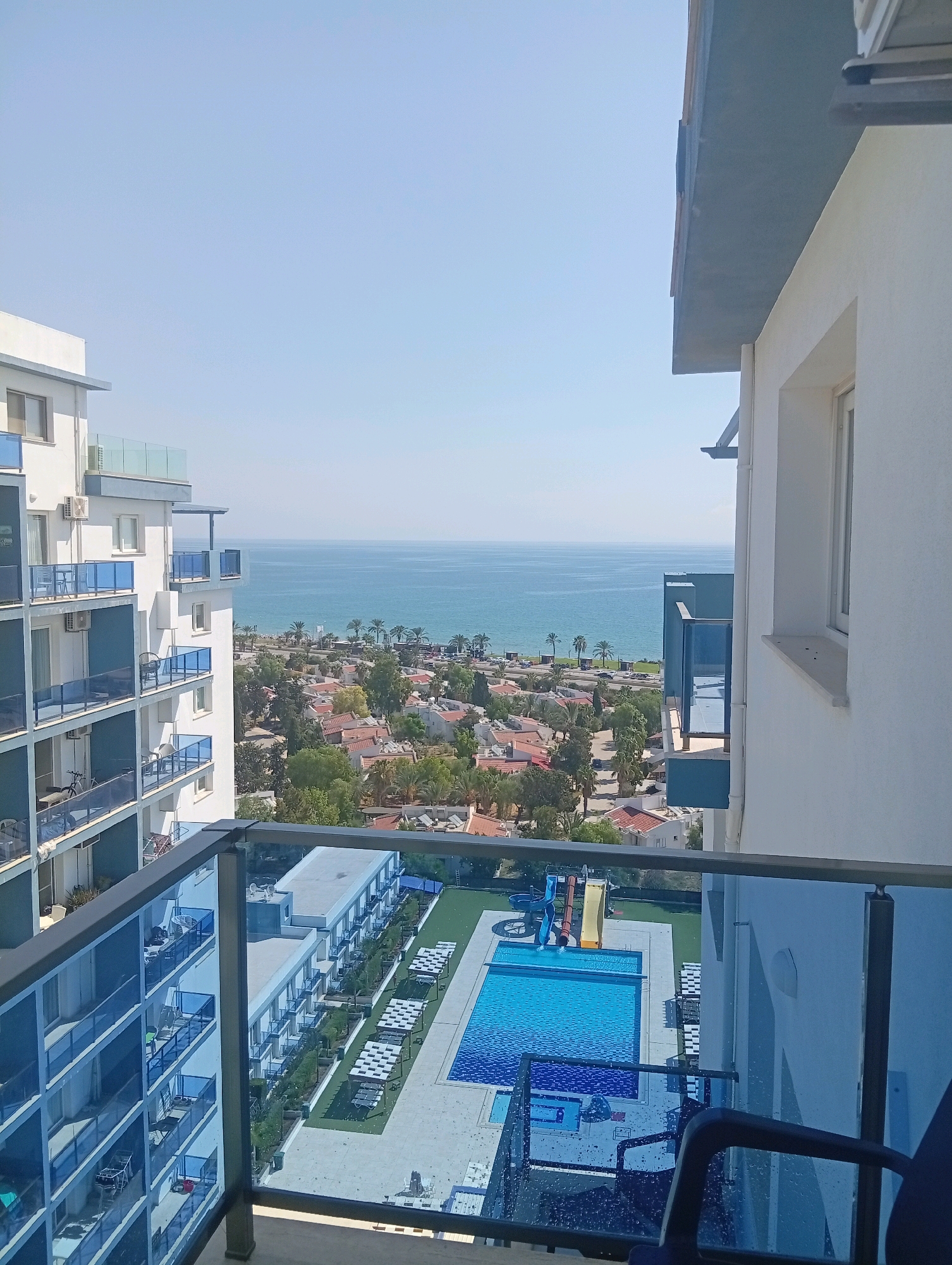This is a high-angle, full shot of a residential complex in Trikomo, Northern Cyprus, on a clear, sunny day. The foreground features a balcony with blue glass railings, overlooking the complex and the sea beyond.
To the left of the frame is a tall, white multi-story apartment building with blue balconies. The building has multiple windows and air conditioning units visible on its facade.
In the middle ground, a courtyard area is visible with a large, rectangular swimming pool with blue water. Adjacent to the pool are children's water slides, a grassy area, and several rows of white lounge chairs and umbrellas. Further back, a mix of smaller residential buildings with red-tiled roofs and trees are scattered, leading towards the coastline.
The background is dominated by the calm, blue expanse of the sea meeting a clear, pale blue sky. Palm trees are dotted along the coastline. The overall scene depicts a resort-like environment. There are no people visible in the image.