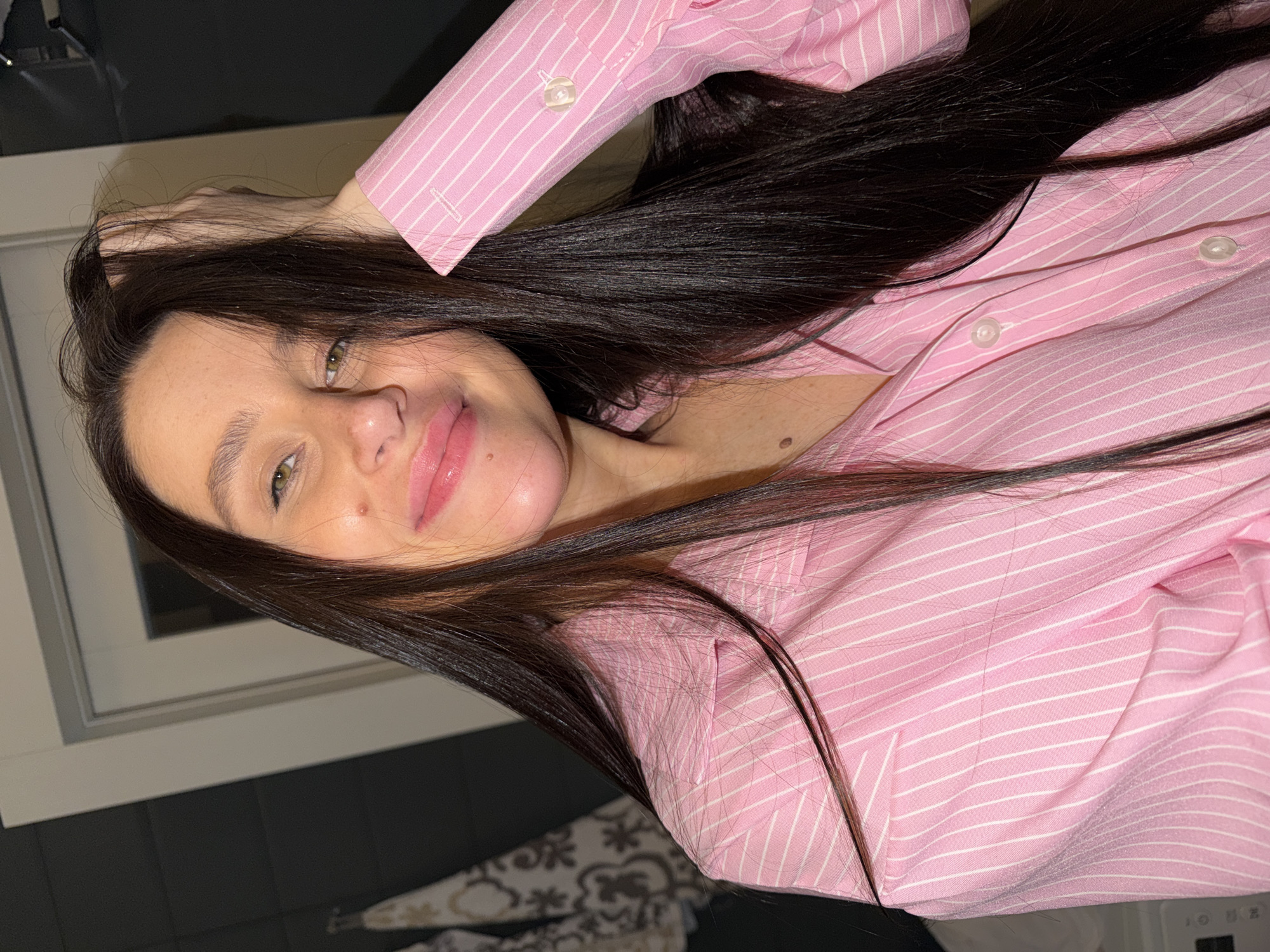 A close-up portrait shows a young woman with long, dark brown hair and striking light-colored eyes, possibly green or hazel. She smiles softly at the camera, her lips coated with a pink gloss or lipstick, conveying a friendly and relaxed expression.
She is dressed in a pink collared shirt with thin white vertical stripes. Her right arm is raised, with her hand positioned near her head, appearing to run through or rest upon her dark hair. Visible on her face are small moles, including one above her upper lip and another near her nose. Additional moles are seen on her neck or upper chest area.
The image is captured indoors from a slightly tilted angle, suggesting she might be lying down or reclining. In the background, dark-colored tiled flooring is visible at the lower left, adjacent to a patterned fabric, potentially a blanket or rug. A white window frame is discernible behind her head, with a dark, indistinct area beyond it, which could be an outdoor view at night or a dark interior space. The lighting is soft and appears to be artificial, illuminating her features and hair.
No discernible text is visible within the image itself. The setting is indoors, and the provided context places the location in Khimki, Russia.
