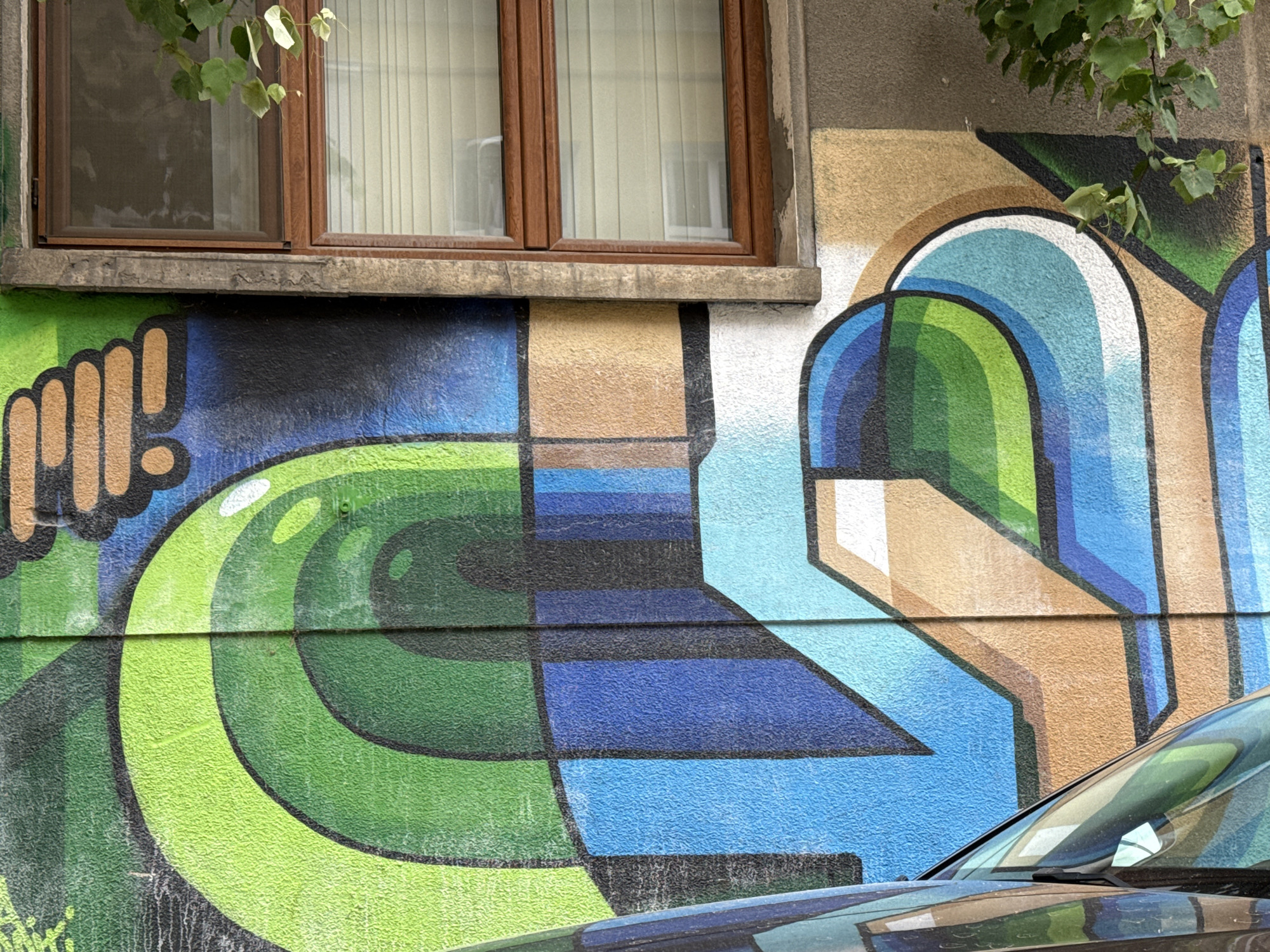 The image shows a graffiti mural on the side of a building in Sofia, Bulgaria. The mural features abstract shapes and bold colors, primarily greens, blues, and browns. A window with wooden frames is visible in the upper part of the image, above the graffiti. The lower portion of the image shows the hood and windshield of a dark-colored car, partially obscuring the lower part of the mural. Green leaves from an overhanging tree are also visible in the top right and left corners of the frame. The wall appears to be textured, and some streaks of dirt or grime are present.
