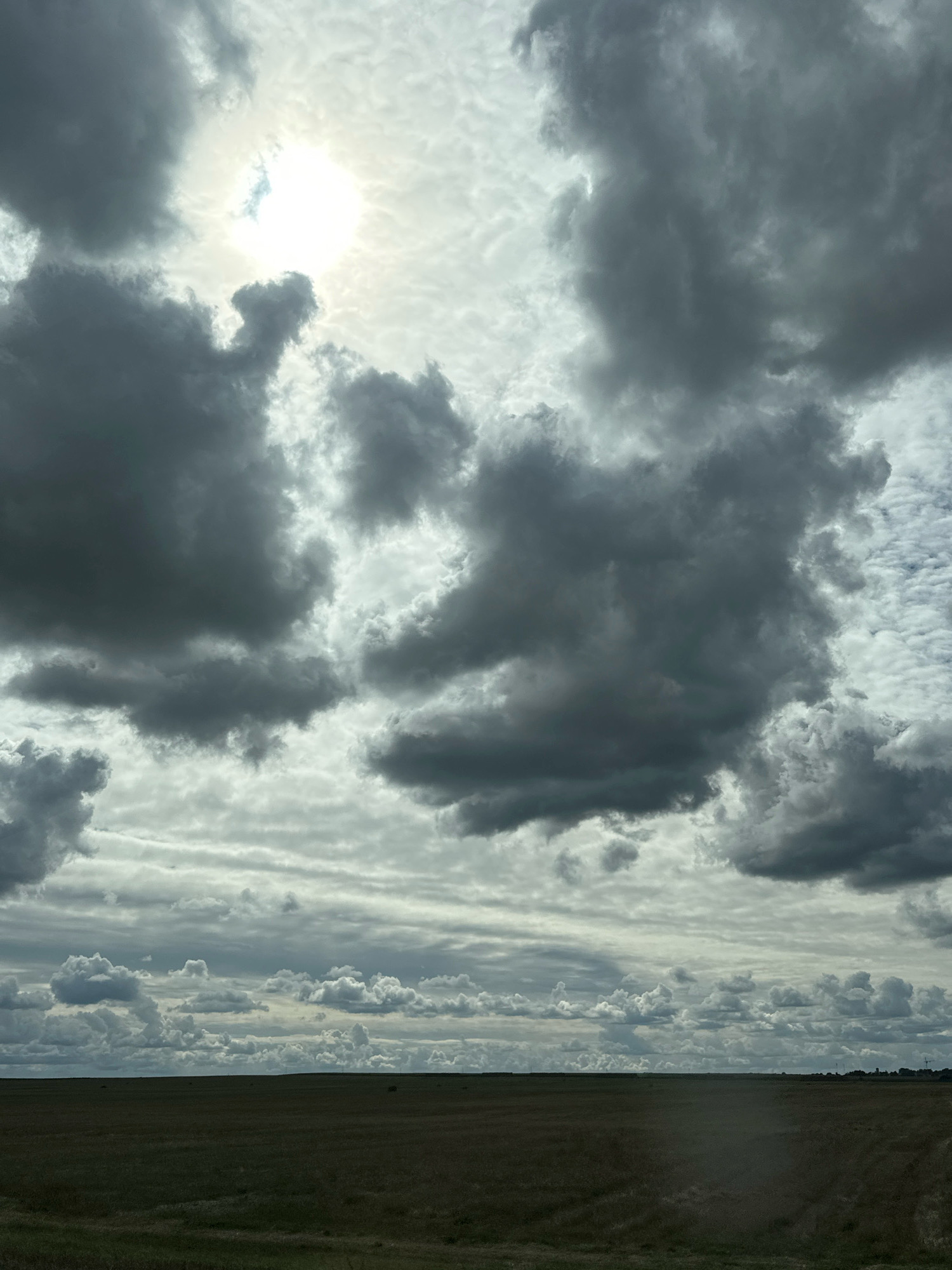 The image depicts a vast, flat landscape under a dramatic, cloudy sky. The foreground consists of a dark, open field covered in grass or low vegetation. The horizon is a sharp, dark line separating the field from the sky. The sky itself is filled with a variety of clouds, ranging from bright, diffused sunlight filtering through lighter, textured clouds to large, dark, heavy storm clouds dominating the upper portion of the frame. The sun is partially obscured by these clouds, creating a bright, luminous area in the upper left quadrant of the image. There are no visible people, structures, or specific landmarks that would indicate a location within the city of Блонскі сельскі Савет, Belarus. The scene is entirely natural, focusing on the interplay of light and shadow in the sky and the expanse of the undeveloped land.
