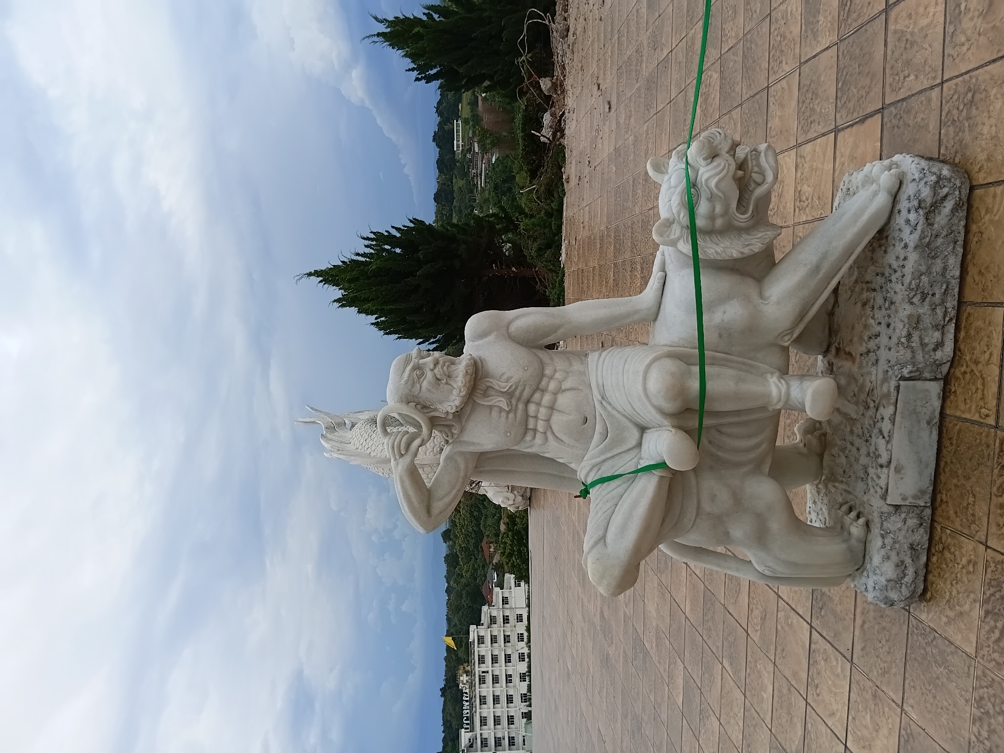 A white marble sculpture of a muscular man with a beard is depicted sitting on a white marble tiger. The man is wearing a loincloth and holding a ring in his right hand, which is raised to his head. A green ribbon is wrapped around the tiger's neck and also around the man's legs. The sculpture is situated outdoors on a tiled patio. In the background, a large white building with several floors is visible, along with lush green trees and distant mountains under a cloudy sky. This scene is located in Ban Lao Phattana, Thailand.
