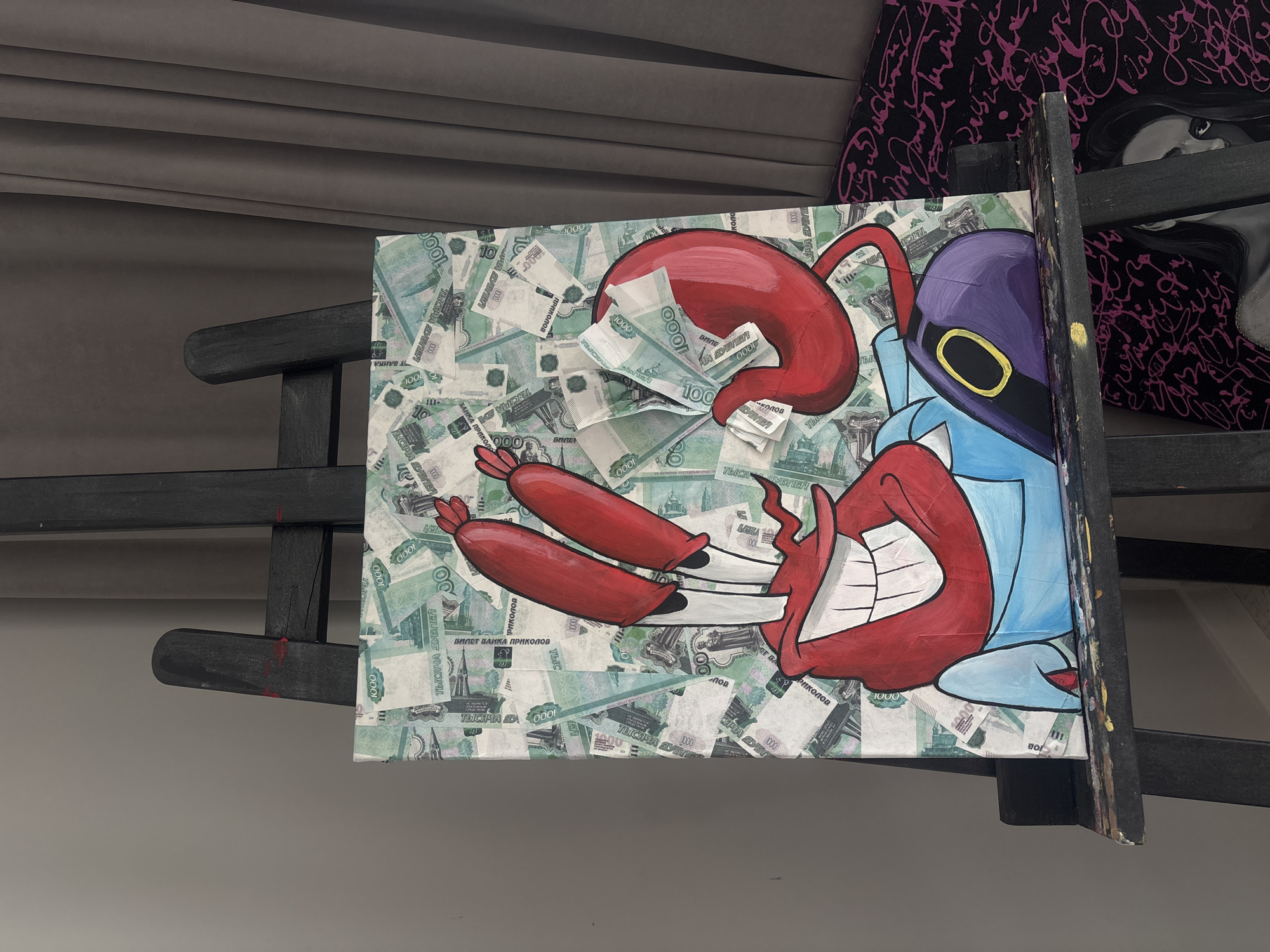 The image shows a painting of Mr. Krabs from Spongebob Squarepants, rendered in a distinctive art style. The background of the painting is composed of what appears to be Russian currency, specifically 1000 ruble notes, arranged in a collage. Mr. Krabs is depicted with his signature red claws and mouth, wearing a light blue shirt and a purple apron with a yellow oval detail. The painting is displayed on a black easel. To the right of the easel, a dark gray curtain hangs. Below the easel, another artwork is visible, featuring a black background with ornate pink calligraphy and a partial image of a woman with dark hair and wide eyes, rendered in grayscale. The overall setting appears to be indoors, possibly an art studio or gallery. The image does not provide any specific visual cues that would allow for the identification of the city of Krasnoyarsk, Russia.