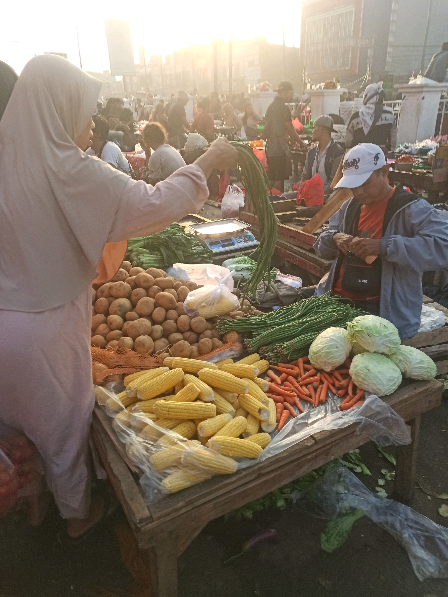 The image depicts an outdoor market scene, likely in Cikarang, Indonesia, given the provided context, with a low-angle perspective and bright, hazy sunlight illuminating the background. In the foreground, a woman wearing a light-colored hijab and a pink garment is standing behind a stall laden with produce. She is holding a bunch of green beans, with her right arm extended. To her right, a man wearing a blue jacket over an orange shirt and a white baseball cap is seen from the chest up, focused on something in his hands.
The stall itself is covered with a variety of fresh vegetables. Piles of potatoes are visible, along with several ears of corn, some wrapped in plastic. Bunches of long green beans and carrots are arranged neatly. Further back, heads of cabbage are stacked, and other green vegetables are present. In the background, a bustling market environment unfolds with numerous people moving about, stalls, and buildings. The overall impression is one of a vibrant morning market.