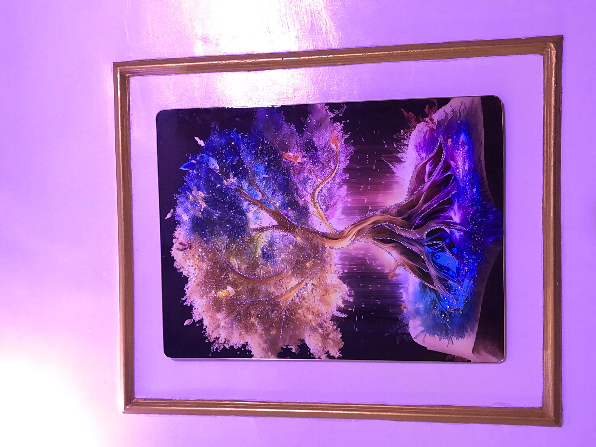 The image captures a framed piece of art displayed on a textured wall, bathed in pervasive purple or magenta ambient lighting. A bright spot of light is visible on the lower left section of the wall.
The artwork itself is a vibrant, stylized depiction of a tree, possibly created with mixed media or digital techniques, featuring a glossy, reflective surface. The central tree structure possesses intricate, gnarled branches and roots, appearing to exhibit a dual nature. The left and central portions of the tree are adorned with warm, golden, and beige-toned foliage, shimmering with numerous tiny, light-colored particles resembling glitter or glowing specks. Small, pale, butterfly-like shapes are also discernible amidst this section.
Contrasting this, the right side of the tree transitions into cooler tones of deep blue and purple, similarly embellished with shimmering particles and subtle dark specks. The background of the artwork is predominantly dark, making the luminous tree elements stand out. Horizontal streaks of light or energy emanate from the right side of the piece, contributing to its dynamic and ethereal quality.
The artwork is housed within a dark, rectangular border, which is then surrounded by a wide, white or off-white mat. This mat is enclosed by a slender, gold-colored frame, creating a layered framing effect. The glossy surfaces of both the artwork and its frame reflect the surrounding purple light. No people or discernible text are present in the image. The scene is static, focusing entirely on the art piece on display.