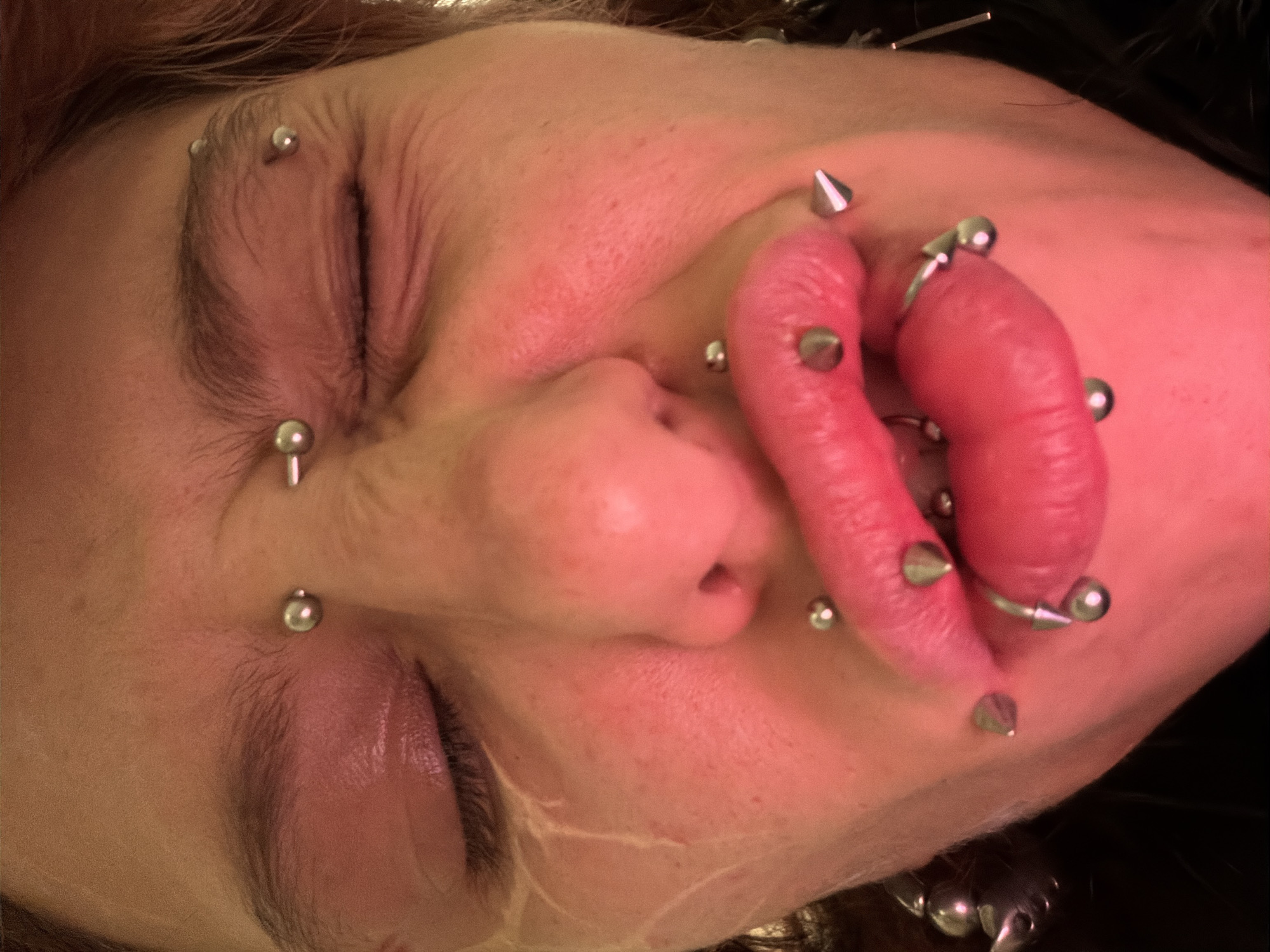 A close-up portrait of a person with numerous facial piercings, including ball-shaped and cone-shaped studs. The piercings are located on the eyebrows, nose, cheeks, and lips. The person's eyes are closed. The background is dark and out of focus. The image provides no clues as to the location in Sofia, Bulgaria.