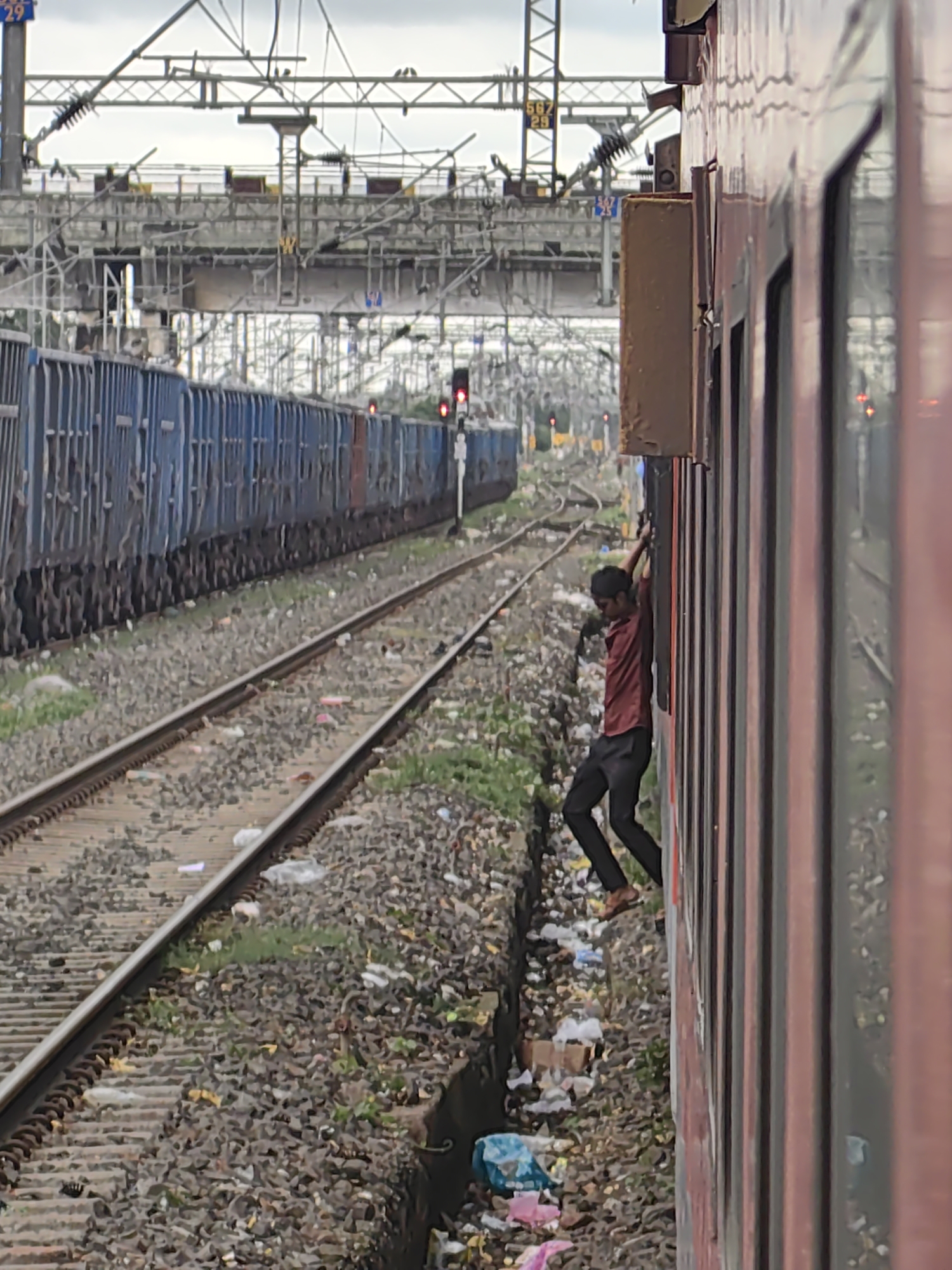 A young man with dark hair, wearing a maroon shirt and black pants, is seen clinging to the side of a red train. His body is partially visible, with his right arm extended upwards, gripping the train's exterior. His legs are bent, and he appears to be in motion as the train travels along the tracks. The train is red with vertical window frames. The surrounding environment consists of train tracks, ballast, and a significant amount of litter and debris scattered along the side of the tracks. In the background, another train with blue carriages is visible, along with overhead electrical lines and railway infrastructure, suggesting a busy railway station or yard. The sky is overcast. The scene is located in Khandwa, India, a city known for its railway junctions and transportation hub.