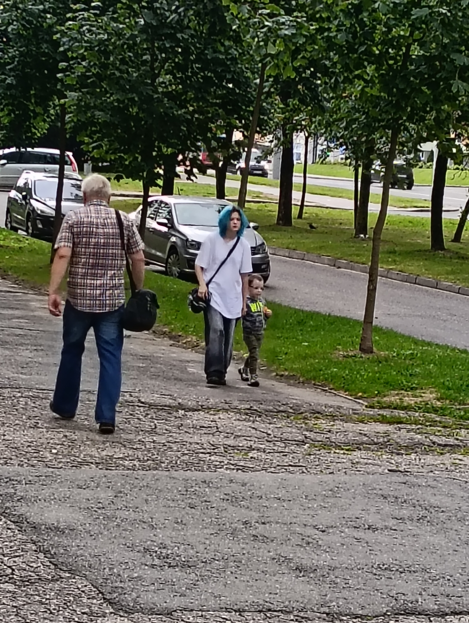 An elderly man with gray hair walks away from the camera, carrying a dark bag over his left shoulder. He is wearing a plaid shirt and blue jeans. To his right, a person with bright blue hair is walking in the same direction, wearing a white t-shirt and loose gray pants, also carrying a bag. A young child in a camouflage outfit walks alongside the blue-haired person, holding something in their right hand. They are on a paved walkway with trees lining the left side and a road with parked cars to the right. The environment appears to be a residential or public area in Hrodna, Belarus, with a focus on the pedestrian movement of the three individuals.
