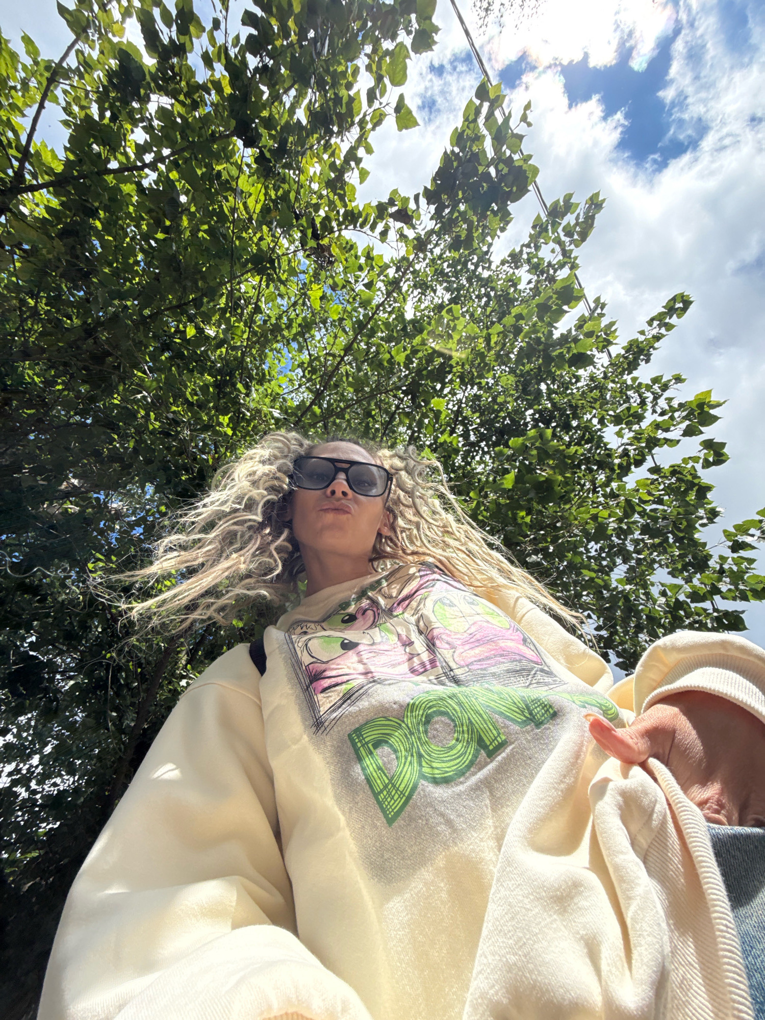 A low-angle, medium shot captures a young woman with blonde, curly hair and large black sunglasses, puckering her lips. She is wearing a cream-colored sweatshirt with a graphic print featuring cartoon characters and the word "DOPE" in green text, layered over a blue denim garment. Her left hand is visible, holding the sweatshirt. She is positioned outdoors, under a large tree with lush green leaves, with a partly cloudy sky visible above. The scene appears to be a candid shot, possibly a selfie. The image does not contain any specific landmarks or elements that would suggest its location is in the city of Rusanovo, Russia.