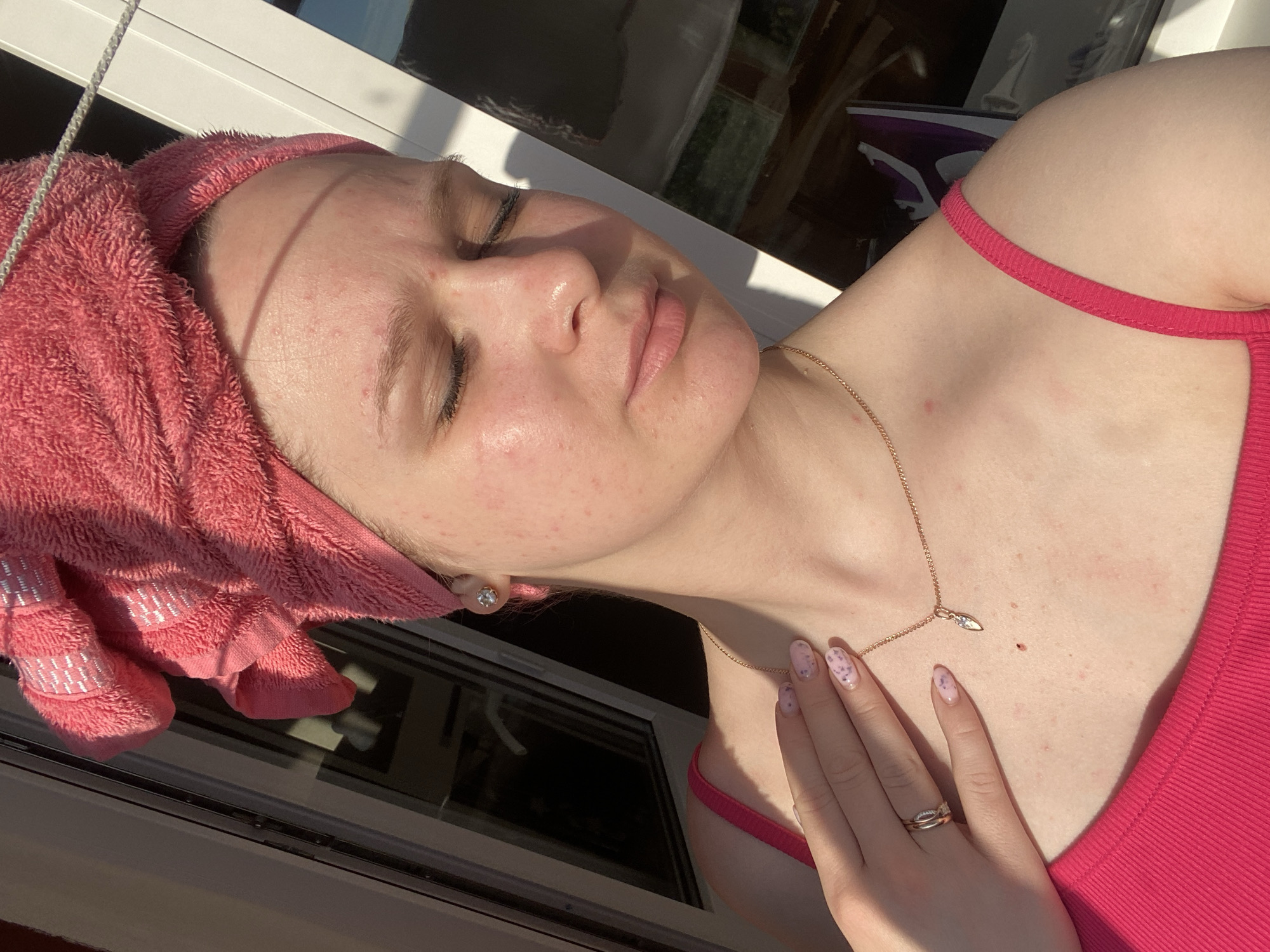 A woman with a pink towel wrapped around her head is positioned near a window, with her face tilted up toward the sunlight. She is wearing a pink tank top and a gold necklace. Her hand is resting on her chest, revealing manicured nails with a speckled design and a ring on her finger. The location is Unknown, Unknown.