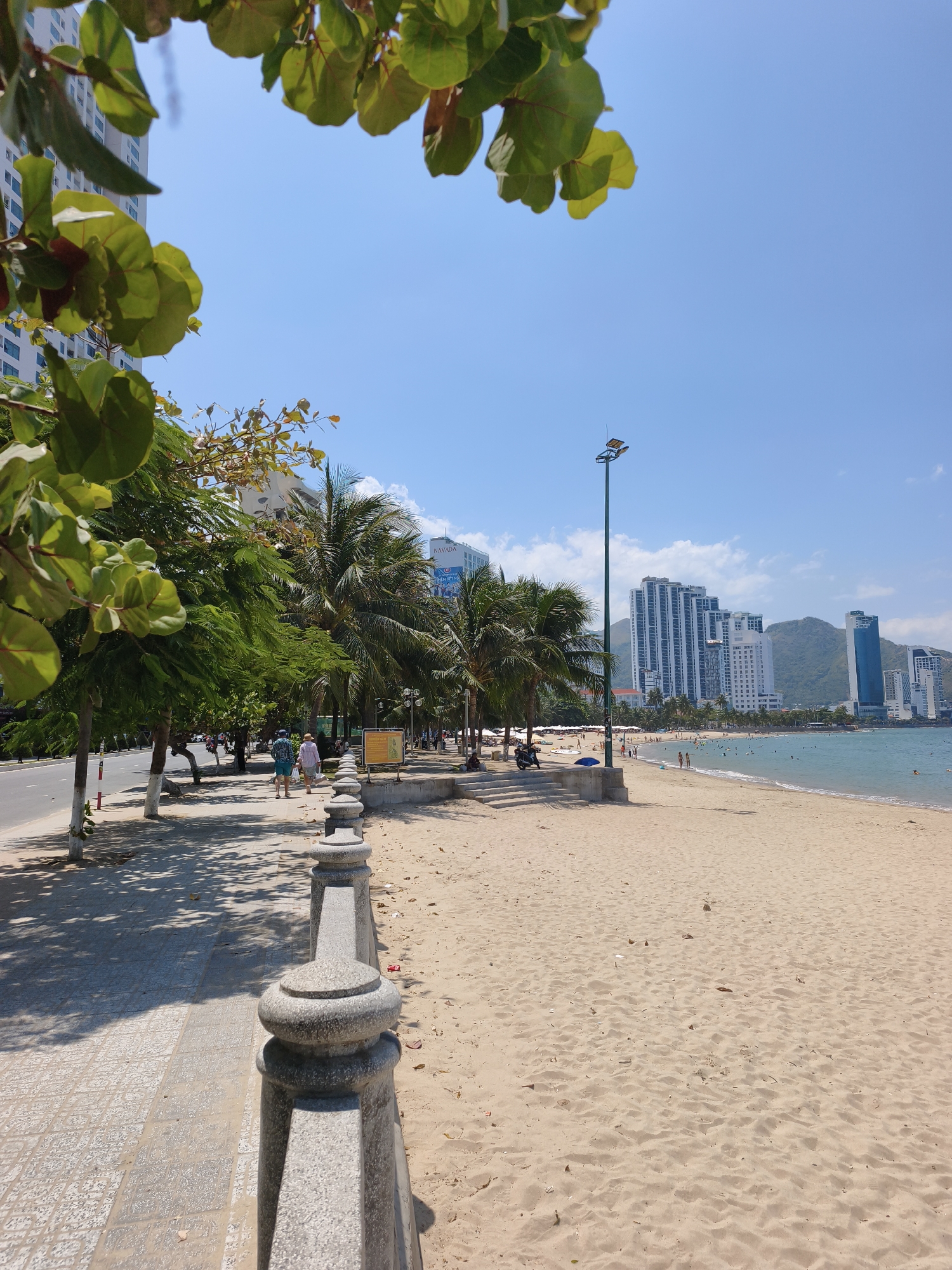 This is a landscape photograph of Nha Trang's beach promenade on a sunny day. The foreground features a sandy beach with a grey stone balustrade running along its edge, separating it from a paved walkway. Beyond the walkway are trees, including palm trees, lining the street. In the background, the ocean is visible, with several people swimming and relaxing on the shore. A row of tall, modern buildings, characteristic of Nha Trang's beachfront architecture, stands behind the trees, with mountains in the far distance. A lamppost is positioned near the center of the image, illuminating the promenade.