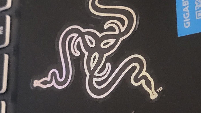 The image displays a close-up, top-down view of a metallic silver and black Razer logo sticker affixed to a matte black textured surface. The sticker features three intertwined snake heads arranged in a triangular pattern. Each snake head is distinctly outlined with pointed snouts and eye-like curves. A small "TM" trademark symbol is visible below the bottom-left snake head. The black surface, consistent with a laptop chassis, occupies the majority of the frame. Portions of three light grey keyboard keys are visible along the top edge, with the letters "ins" partially discernible on one key. A segment of a bright blue sticker with white capital letters reading "GIGABY" is visible in the bottom-right corner. The scene is situated in Magaluf, Spain. No individuals or active interactions are present.