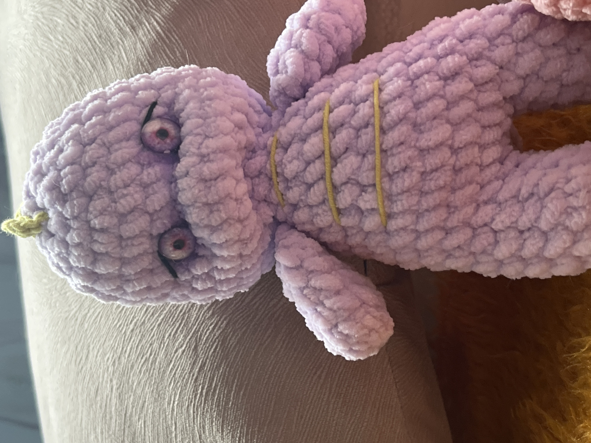 A close-up, high-angle shot shows a light purple crocheted stuffed toy. The toy has large, round eyes with dark pupils and pink irises, outlined by thin black lines. A small yellow tuft of yarn sprouts from the top of its head. Three thin yellow yarn stripes are visible across its torso. The toy is resting on a beige textured surface, with a yellow furry material visible below and to the left. The background is blurred, suggesting a shallow depth of field. The specific location of Khostinskiy, Russia is not evident in the image.