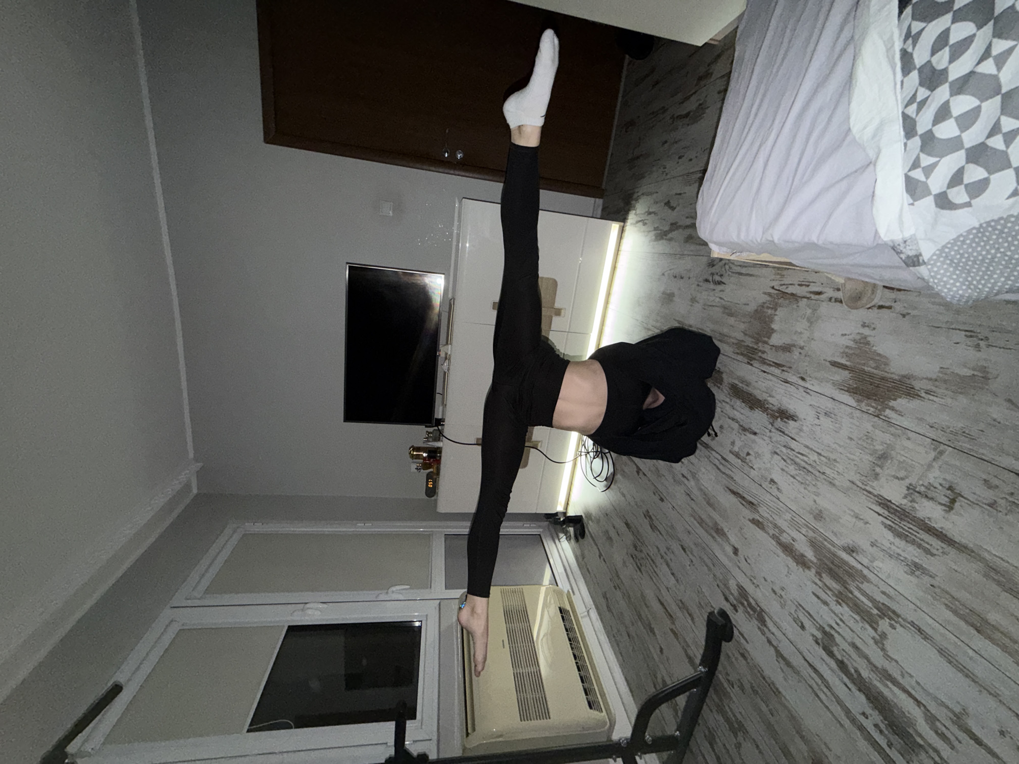 A person in black leggings is performing a vertical split in a room with light-colored wooden floors. The person's right leg, clad in a white sock, is pointing upwards, while the left leg is extended downward towards a window and an air conditioning unit. Behind the person is a white cabinet, a television, and a dark brown cabinet above. A bed with a white and grey patterned comforter is to the right. The location is a private indoor space, not identifiable as specifically Plovdiv, Bulgaria based on visual information.