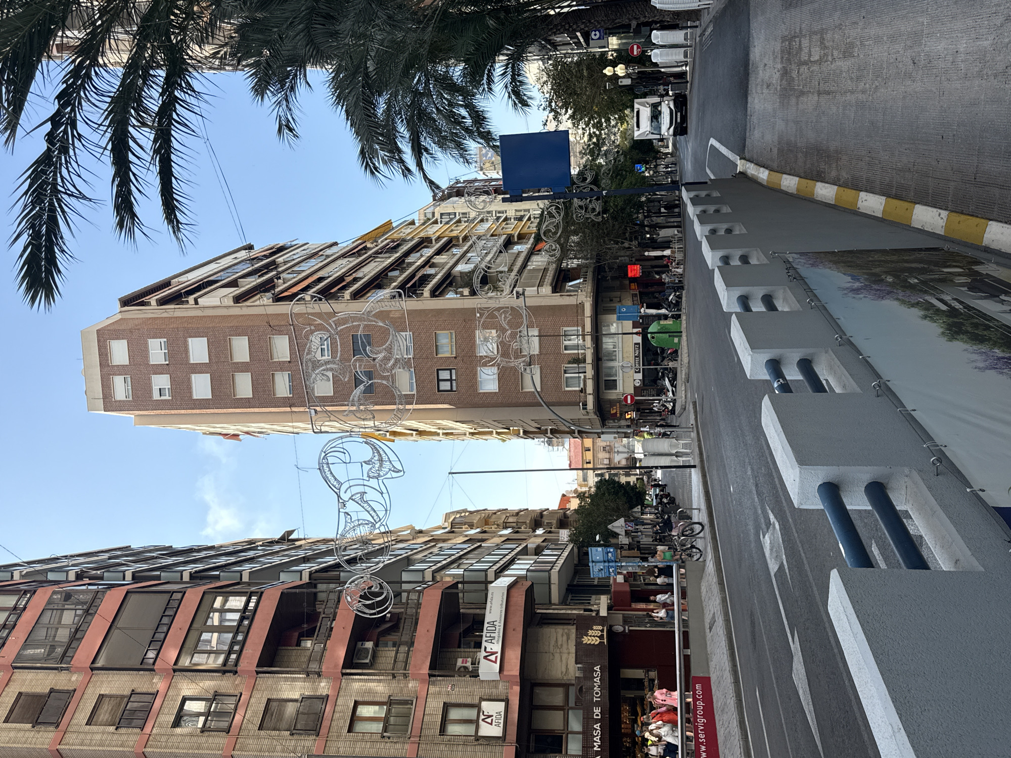 The image depicts a wide urban street flanked by multi-story buildings under a clear blue sky. On the left, several buildings feature reddish-brown brick facades with balconies, some enclosed. At street level, "LA MASA DE TOMASA" bakery and "AFIDA Abogados & Asesores tributarios" are visible. Numerous pedestrians walk on the sidewalk, some gathered near the bakery, while a few sit at outdoor cafe tables. Parked motorcycles and bicycles are present. Elaborate, white metallic scrollwork decorations are suspended across the street.
On the right side, a tall, slender building with a reddish-brown brick facade and white windows is prominent, partially obscured by several large palm trees. Further down the street, a white delivery truck drives towards the viewer. The sidewalk on the right features stacked white plastic chairs and a blue rectangular sign on a pole.
In the foreground, a grey concrete barrier with blue horizontal bars and a banner displaying a nature scene is on the left. The asphalt road has white lane markings, including a large arrow indicating a straight or right turn. A yellow and white striped curb separates the road from a paved area on the right. The overall scene suggests a busy city street in a warm climate, possibly during a festive period due to the decorations.