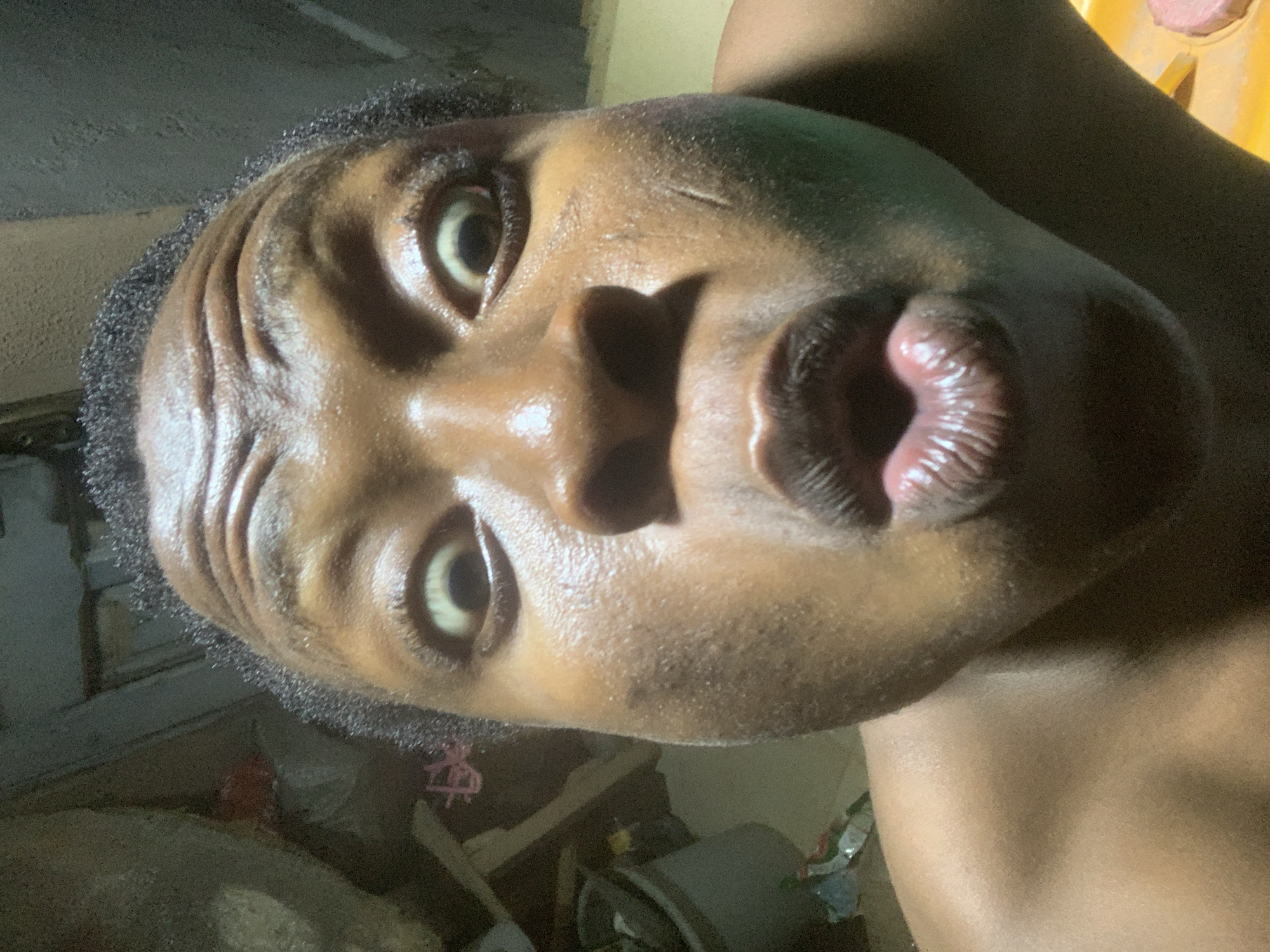 A close-up, tilted portrait shows a person with dark skin, shiny skin on their face, and dark, short hair. Their eyes are wide open, and their lips are pursed. The background is indistinct, suggesting an interior space. The location is Unknown, Unknown.