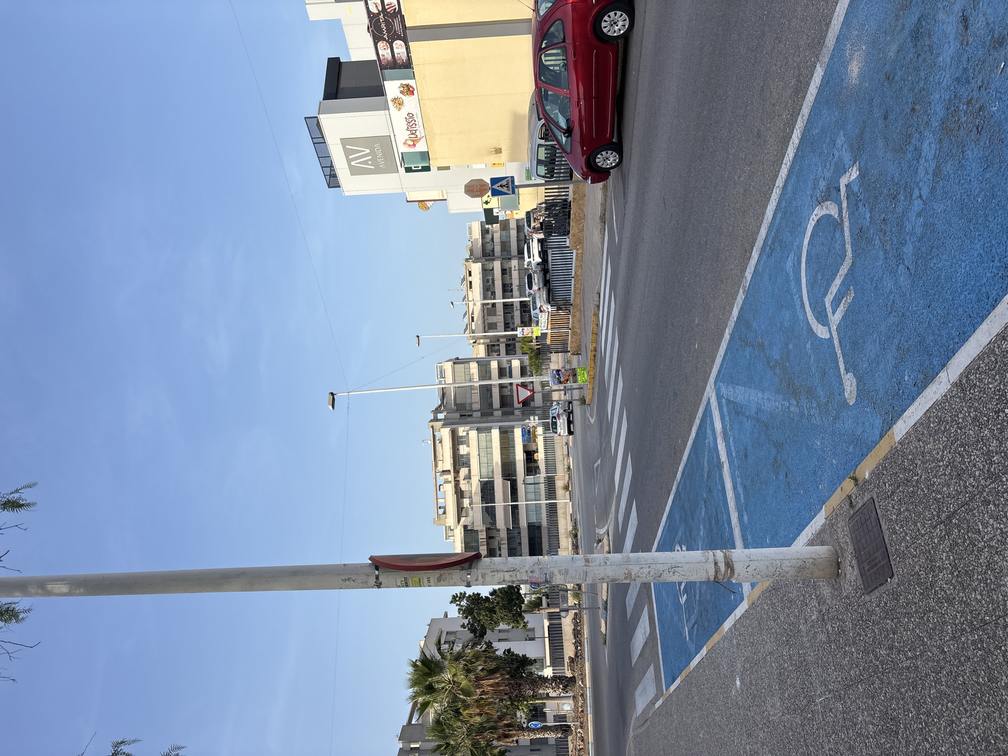 The image shows a street scene in Orihuela, Spain, under a clear blue sky. In the foreground, a lamppost stands on a sidewalk made of dark grey aggregate. Adjacent to the sidewalk is a road with white pedestrian crosswalk markings. A section of the road, marked with blue paint and white lines, designates a parking spot for disabled individuals, featuring the international symbol of access. A red car is parked in this designated spot.
Further down the street, several multi-story apartment buildings are visible, characterized by their beige and white facades and numerous balconies. Palm trees line the sidewalk. Across the street, there's a building with "AV AVENIDA" signage. Other businesses, including one with "Delfissio" and "Amrita" visible, are also present. Traffic signs, including a stop sign, are placed along the road. Several other vehicles are parked or moving in the background. The overall impression is a typical urban streetscape in daylight.