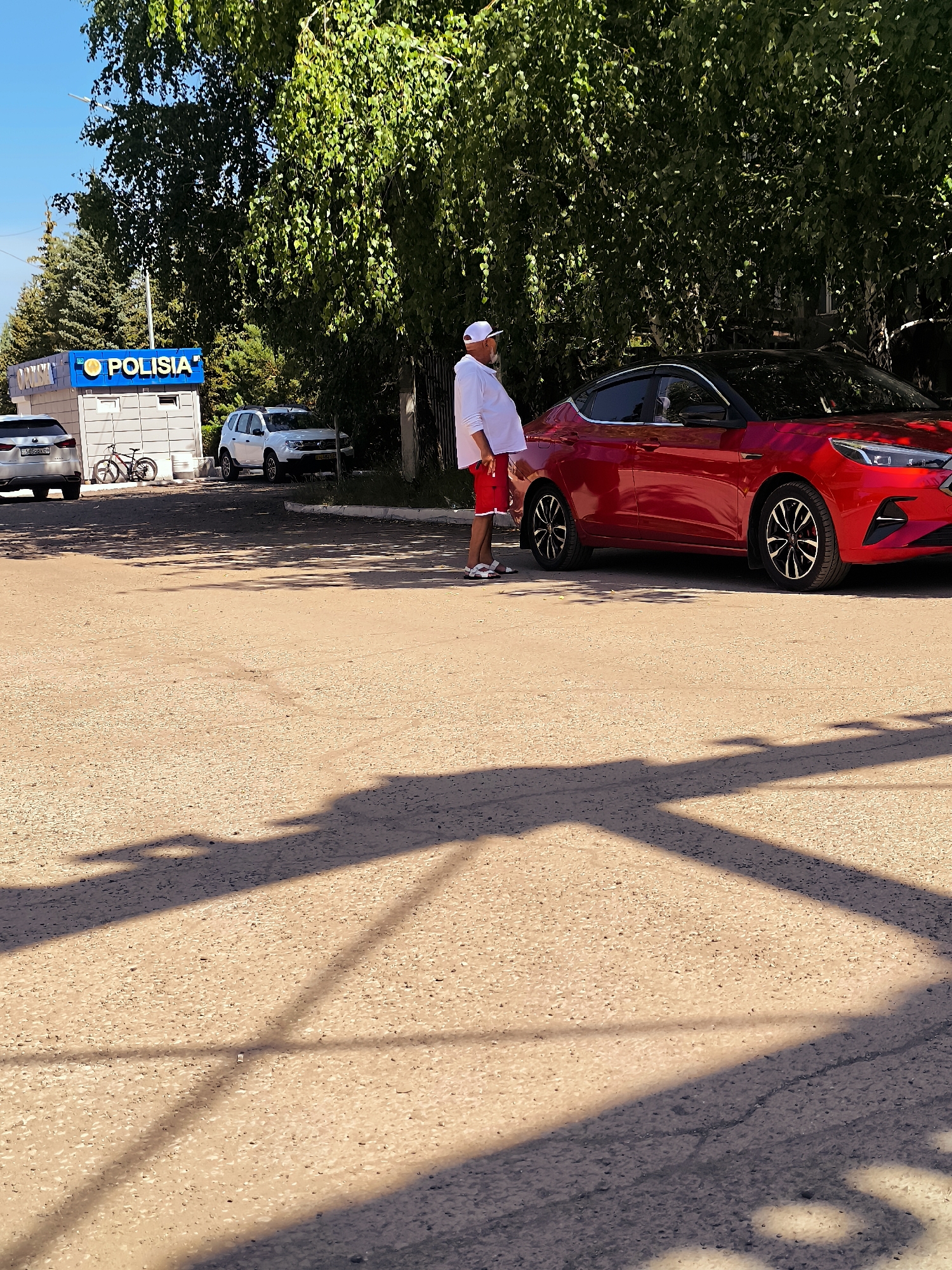 A man wearing a white cap, white long-sleeved shirt, and red shorts stands next to a red car. He is positioned on the right side of the image, facing left, with his right arm bent and resting on his hip. The car is a modern sedan with black accents on the wheels and windows. Behind the man and car, trees form a dense green canopy against a clear blue sky. To the left of the man, a small building with "POLISIA" written in blue letters on its facade is visible, with two cars and a bicycle parked in front of it. The ground is a gravelly, light-colored surface. Shadows of tree branches are cast across the ground in the foreground, indicating a sunny day. The overall scene suggests an outdoor location in a city, possibly a police station or a similar administrative building.