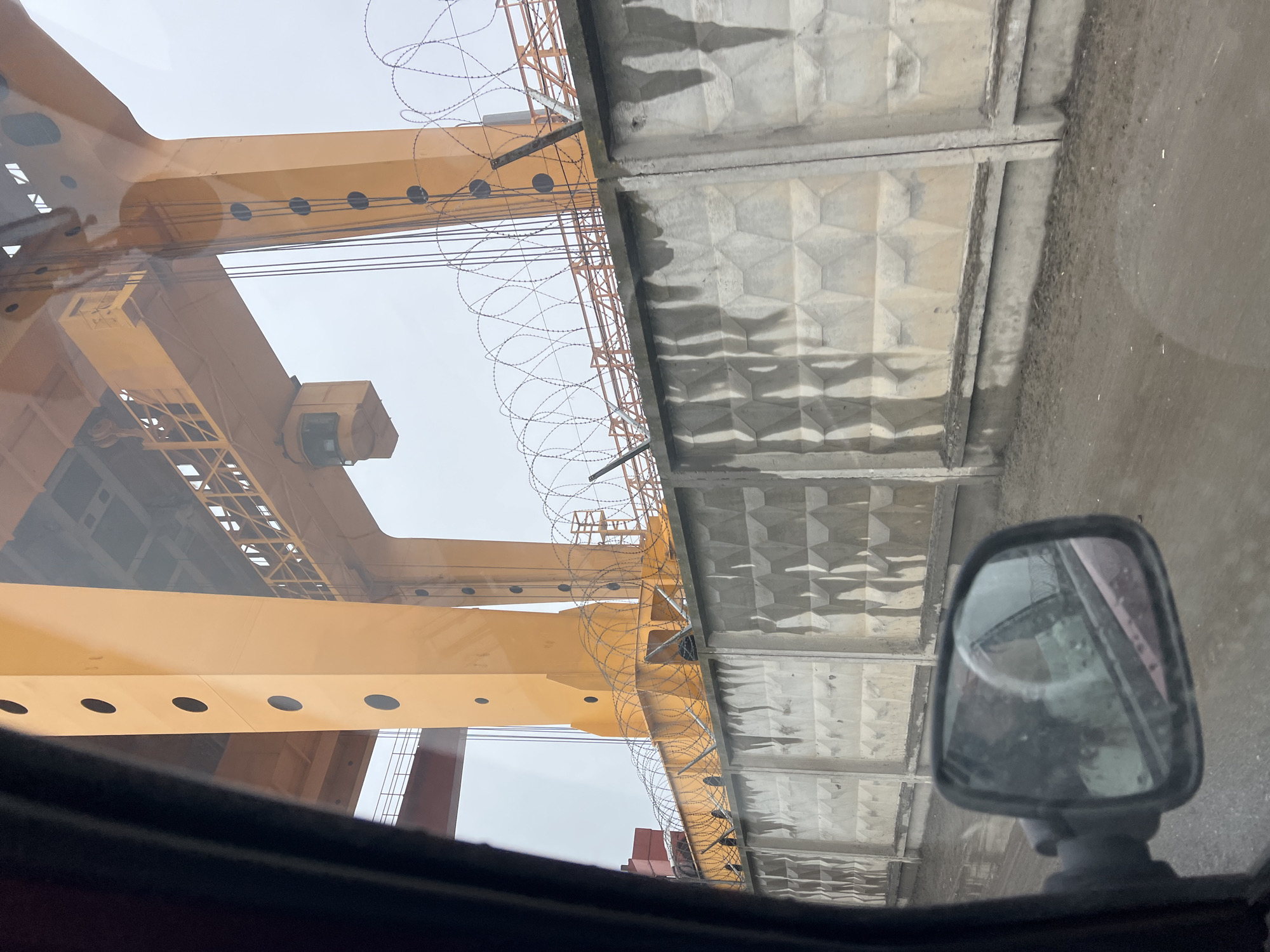 The image, taken from inside a vehicle, shows an industrial scene. A large yellow crane structure dominates the left side of the frame, while a concrete wall runs along the right side. Barbed wire is strung along the top of the wall and parts of the crane structure. A side mirror of the vehicle is visible in the lower right corner of the image, reflecting parts of the scene. The sky is overcast. The location is Noviy, Russia.