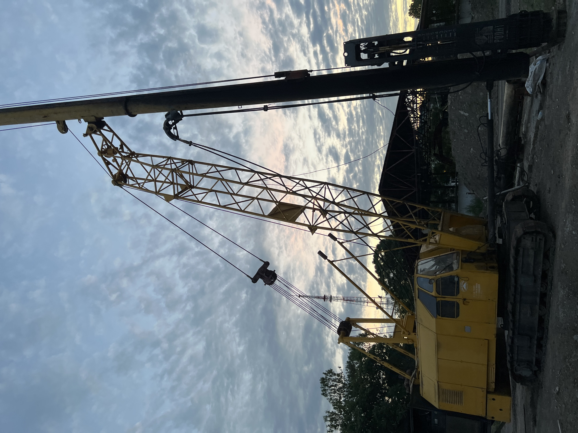The image shows a yellow crawler crane parked on a dirt surface, likely a construction site, in Krasnodar, Russia. The crane is the primary object, with its long boom extending upwards and to the left, partially silhouetted against a cloudy sky. The sky is filled with wispy clouds, and the light suggests it is either early morning or late evening, with a soft glow visible on the horizon.
Notable details include the intricate lattice structure of the crane's boom, the cables, and the pulleys used to operate it. The cab of the crane is visible, with windows and doors, appearing to be unoccupied. The crawler tracks of the crane are also visible, indicating its mobility. In the background, there are hints of industrial structures, possibly a building under construction or a factory, and trees. A pile of dirt or gravel is also present.
On the vertical structure to the right of the crane's boom, there is visible text. It includes a logo with a bear-like figure and the word "PORAT" in capital letters, followed by smaller text below that is too blurry to read. There are also numerical markings on this vertical structure. The overall mood of the image is quiet and still, suggesting the end of a workday or the beginning of one, with the equipment at rest. The lighting creates a somewhat dramatic and serene atmosphere.