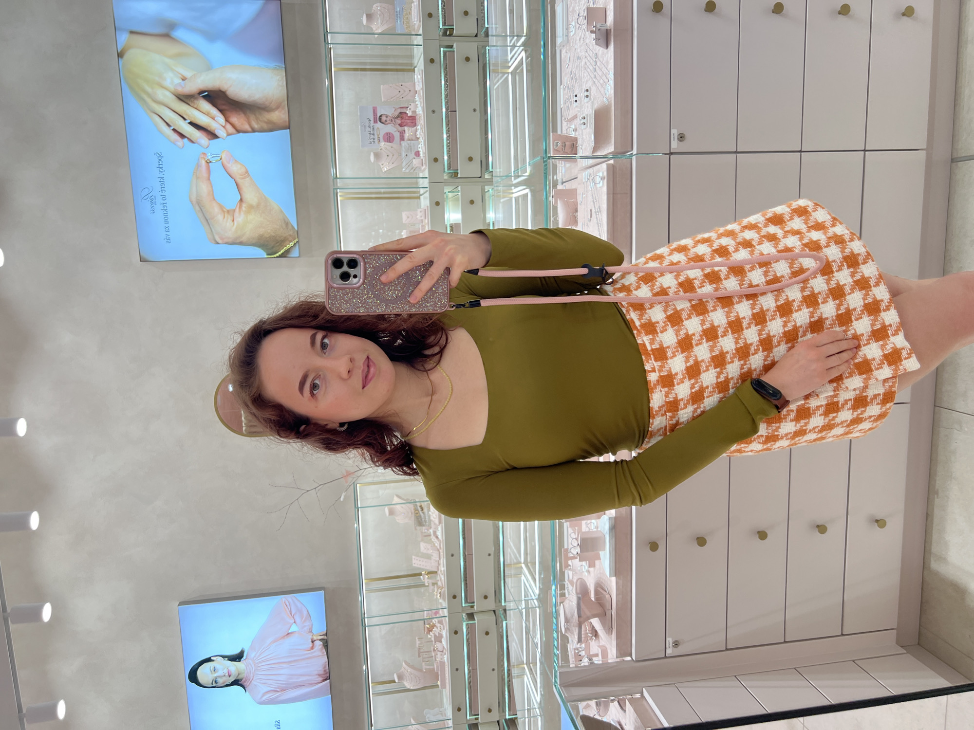 A woman with curly brown hair, wearing a green long-sleeved top and an orange and white checkered skirt, takes a selfie in a jewelry store. She is holding a pink glitter-covered phone with a pink strap. Behind her, glass display cases filled with jewelry are visible. Two large screens are mounted on the wall above the display cases; one shows a woman in a pink blouse, and the other shows hands trying on a ring. The store appears clean and well-lit.