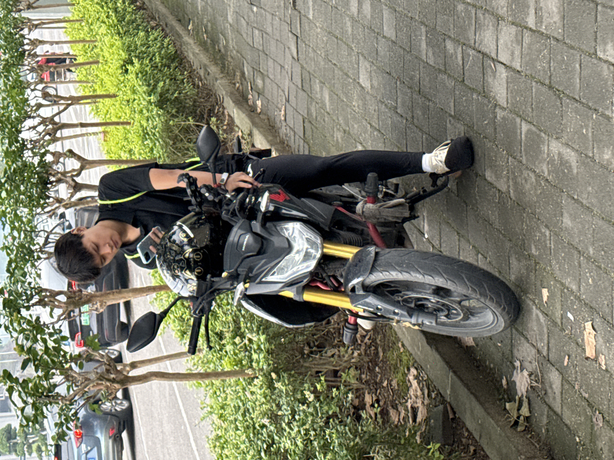A young person is shown in an outdoor, daytime setting, seated on a black and grey Honda motorcycle with distinctive gold-colored front forks. The motorcycle is stationary on a paved area, likely a sidewalk or road shoulder. The person, who appears to be male, wears a black short-sleeved top with a neon green accent, black athletic pants, and white and black sneakers. A smartwatch is visible on their left wrist. They are holding a light-colored smartphone in their left hand and looking down at it, appearing engrossed. A black helmet with a gold patterned design rests on the motorcycle's handlebars.
The ground consists of grey rectangular pavers. To the person's left, a row of green bushes and small, sparsely leaved trees line the paved area, with parked cars visible on an asphalt road in the background. To the right, a low concrete curb separates the pavers from a patch of dirt with fallen leaves. The lighting suggests an overcast or partly cloudy daytime, casting soft, even illumination on the scene. The visible text "HONDA" is present on the motorcycle's tank area.