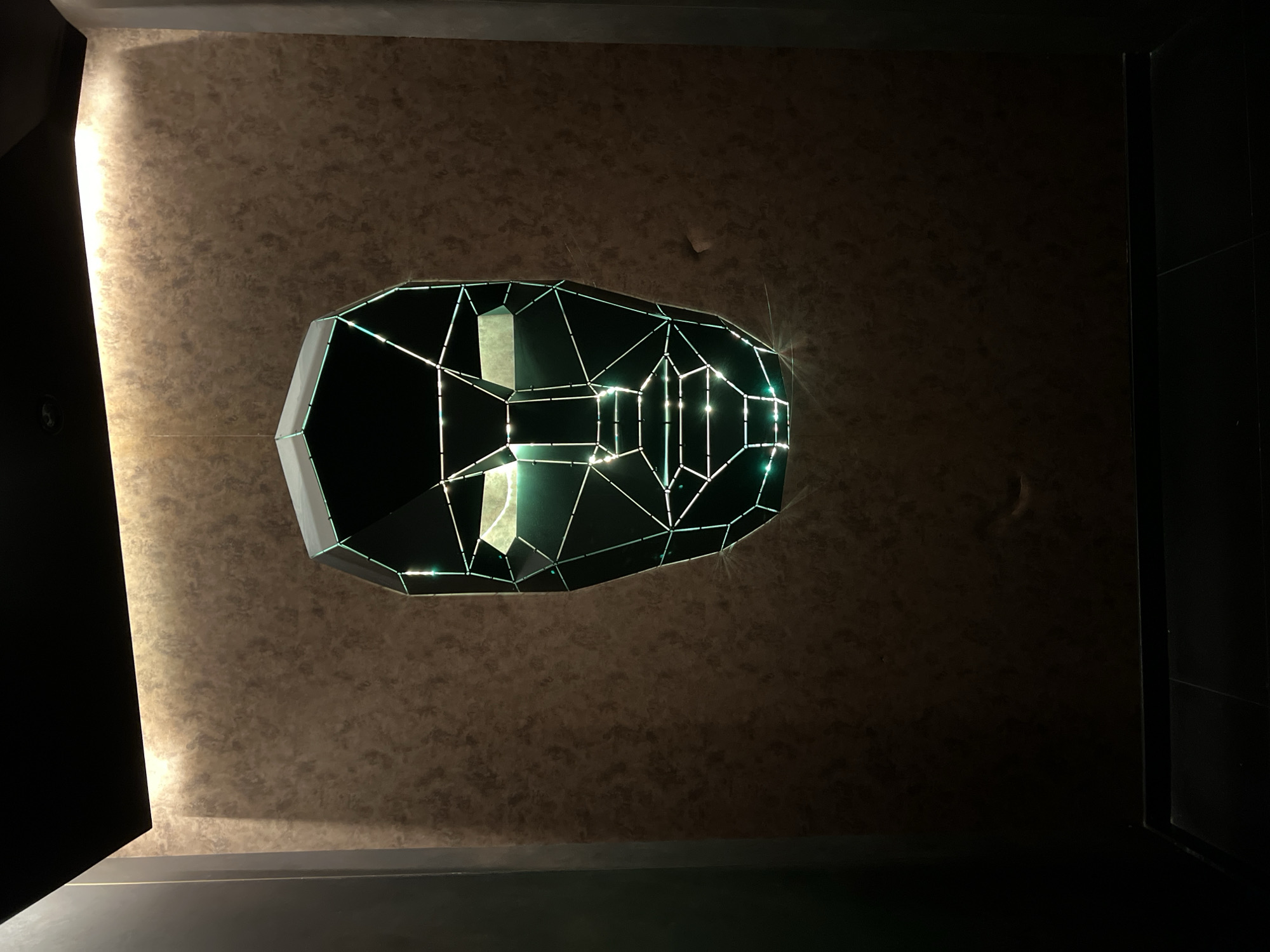The image depicts the ceiling of an interior space. Suspended from the ceiling is a geometric, polyhedron-shaped light fixture. The ceiling has a mottled, textured appearance, possibly wallpaper or a decorative paint finish. A vertical, dark edge is visible on the right side of the image. The location is most likely Burgas, Bulgaria. No people or actions are visible.