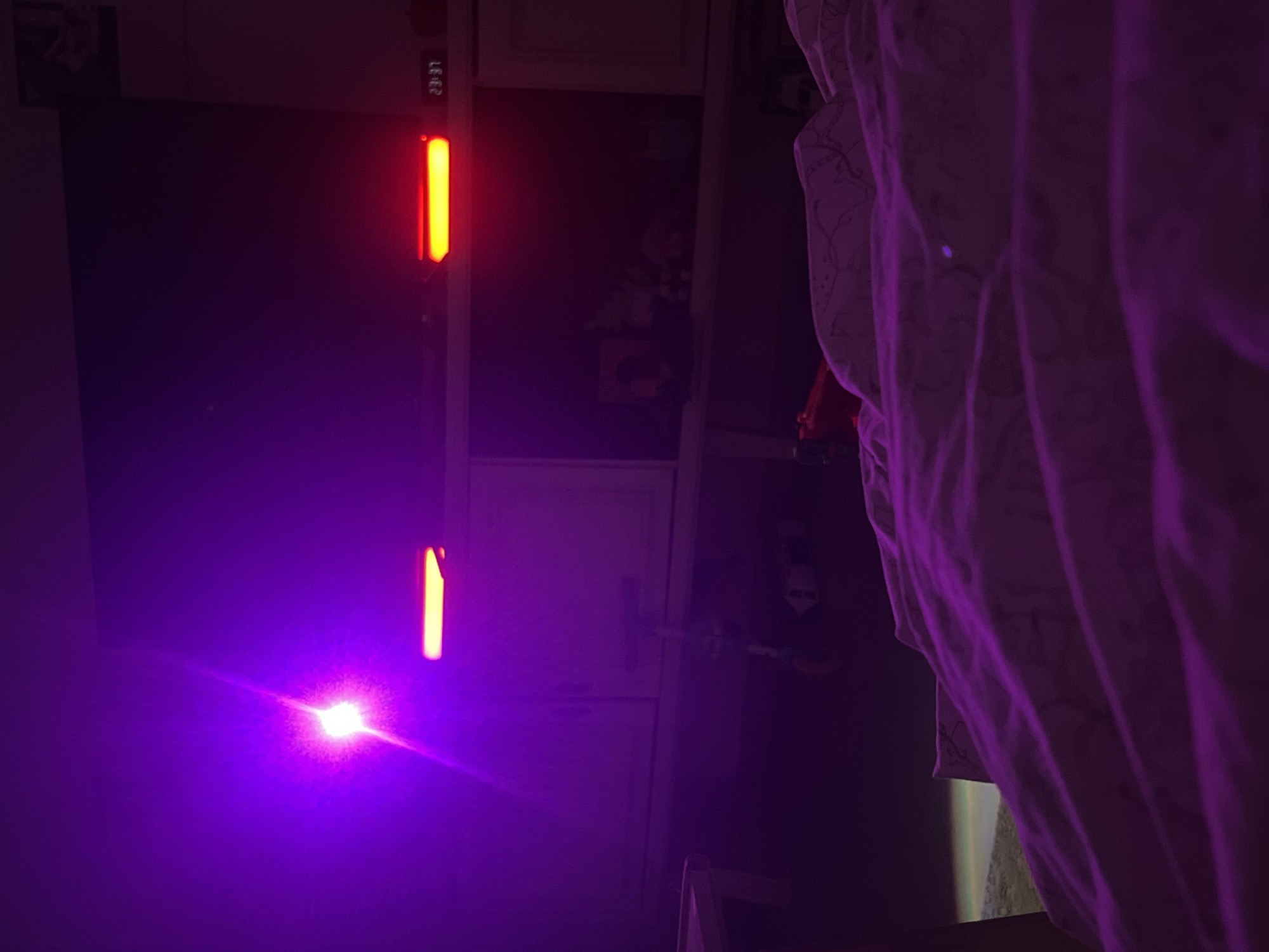 The image shows a room with a TV screen displaying an intense purple light and reflections. Next to the TV is a vertical LED light strip, showing '23:31' and a red/orange color. The room appears to be a bedroom due to the presence of a patterned blanket/sheet on the right side. The backdrop appears to be kitchen cabinets. Location information is not discernible from the image, and location within Kiryat Bialik, Israel, cannot be determined.