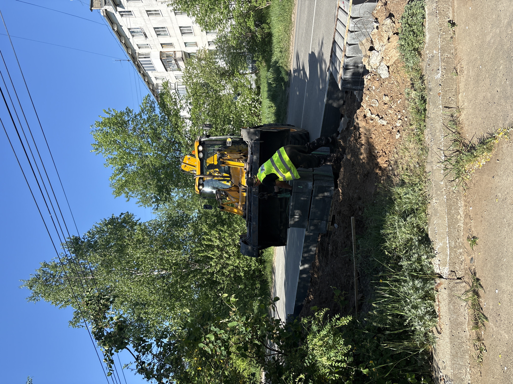 The scene shows a construction site next to a road. A yellow backhoe is positioned near a partially excavated trench. A worker wearing a bright yellow safety vest is bent over in the trench, possibly working on the installation of materials. Gray paving stones are stacked along the edge of the trench. Vegetation, including grass and shrubs, grows along the roadside. In the background, there is a multi-story apartment building with white walls and numerous windows. Power lines are visible in the upper left corner of the image. The sky is blue, suggesting a sunny day. The scene suggests ongoing infrastructure or road construction. The image appears to be taken in Chaikovsky, Russia, as indicated in the instructions.