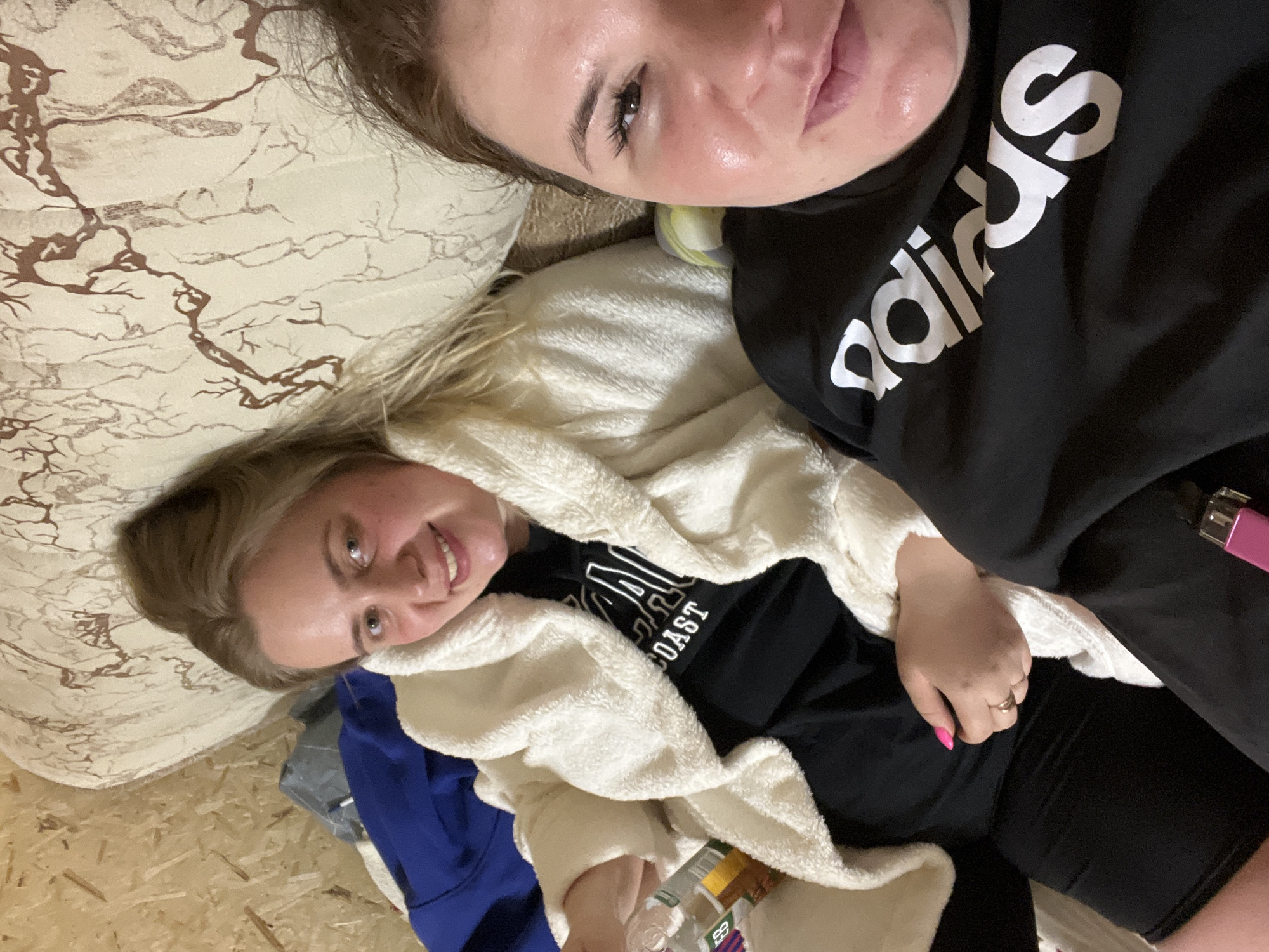 Two women are lying down indoors. One woman, with blonde hair, is covered in a white fluffy robe, wearing a black t-shirt with "COAST" written on it. She is holding a plastic bottle. The other woman is wearing a black Adidas sweatshirt. They appear to be on a bed with a patterned beige sheet, and the floor is made of wood planks. The location is potentially a private residence, but the available information doesn't contain location details for Chaikovsky, Russia.