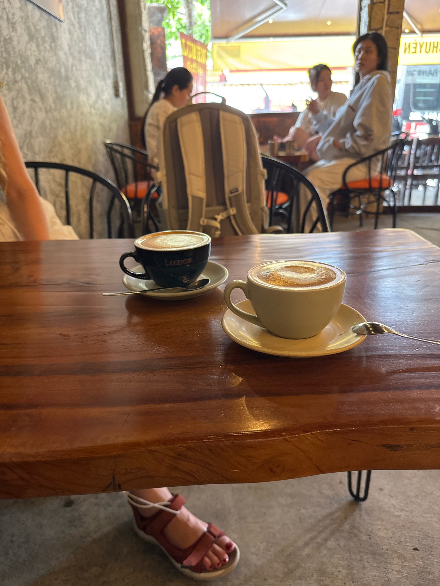 The image captures a close-up view of a wooden table at a cafe in Nha Trang, Vietnam. Two cups of coffee are prominently displayed on the table, one in a black cup with a gold logo reading "Lumnen" and the other in a beige cup. Both cups are saucers and have spoons resting on them. The coffee in both cups appears to be prepared with latte art. In the background, out of focus, several people are seated at other tables, suggesting a social gathering. The cafe's ambiance is casual and relaxed. The lighting suggests it is daytime, and the overall scene evokes a sense of everyday life in a Vietnamese city.