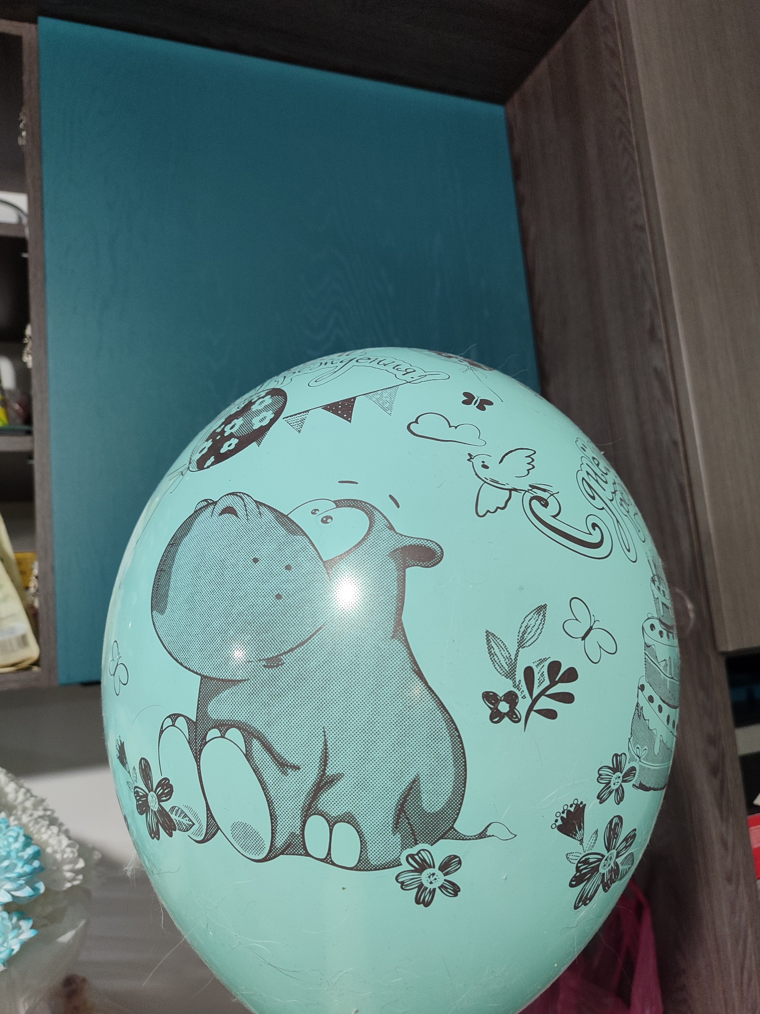 A light blue (aqua/teal) balloon, prominently decorated with cartoon-style illustrations in dark gray, is the central focus of the image. A large, smiling hippopotamus, depicted sitting with its feet forward, is the main illustration on the balloon's front. Surrounding the hippopotamus and across the balloon's surface are various festive and natural elements: butterflies, assorted flowers with leaves, clouds, a bird carrying a banner, a string of pennant flags, another smaller balloon with a polka-dot pattern, and a multi-tiered birthday cake. The banner carried by the bird clearly displays the Russian text "С днём рождения!" (Happy Birthday!).
The balloon is positioned indoors, likely in a room in Izhevsk, Russia, with visible background elements suggesting furniture and decor. To the left, a flat, dark teal-colored panel or surface is visible, possibly part of a cabinet or wall accent. To the right and top, dark brown wooden cabinetry with a visible grain provides a warm contrast. In the bottom left corner, blurred white and light blue floral decorations, possibly artificial flowers or fabric, are partially visible. The overall scene suggests a celebratory atmosphere, likely for a birthday event. The lighting is bright and even, indicating an indoor setting during the daytime or with good artificial illumination.
