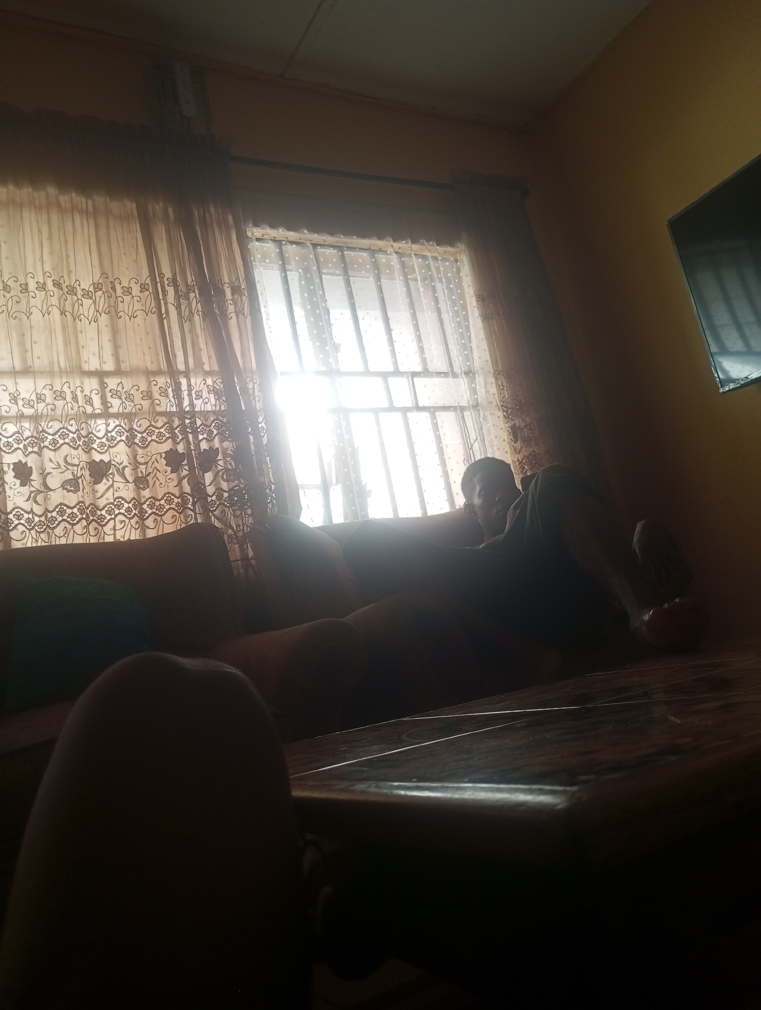 In a dimly lit room in Onitsha, Nigeria, a person is lounging on a sofa, facing away from a window. The window is covered by sheer, patterned curtains, and outside, sunlight is filtering through the glass. A television is mounted on the wall to the right of the window. In the foreground, a dark, polished surface, possibly a table, is visible. The scene appears to be a domestic indoor setting.
