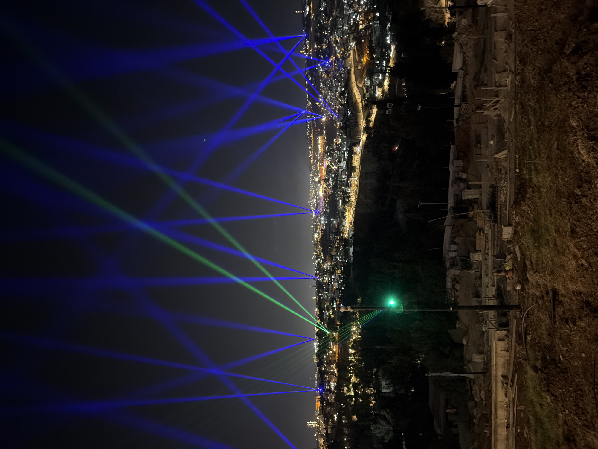 The photograph captures a night view of Jerusalem, Israel, with numerous blue and green laser beams projected into the dark sky from various points within the city. The lasers create an impressive light show, reaching high above the urban landscape. Below the lasers, the city lights are visible, spread across hills and valleys, indicating a densely populated area. The foreground features a construction site with piles of dirt, debris, and building materials, suggesting ongoing development or renovation. A single green traffic light is illuminated on a pole in the foreground, adding a touch of urban infrastructure to the scene. The overall impression is one of a vibrant city illuminated by a dramatic light display against the backdrop of night.