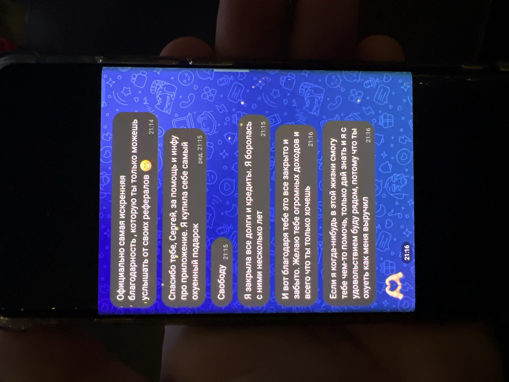 Here is a description of the image:
**Scene:** The image depicts a smartphone screen displaying a Telegram chat. The background of the chat is a blue pattern with various images. The chat messages are written in Russian.
**Content of Chat Messages (Translated):**
* **Message 1:** "Officially, the most sincere gratitude you can only hear from your referrals."
* **Message 2:** "Thank you, Sergey, for the help and info about the application. I bought myself the most amazing gift."
* **Message 3:** "Freedom."
* **Message 4:** "I have closed all debts and loans. I fought them for several years."
* **Message 5:** "And now, thanks to you, it's all closed and forgotten. I wish you huge incomes and everything you want."
* **Message 6:** "If I can ever help you with anything in this life, just let me know, and I will be happy to be there for you because you helped me out immensely."
**Other details:** The phone is held in someone's hand, with fingers partially visible. The lighting is somewhat dim, indicating a low-light environment.
**In summary:** The image shows a Telegram chat where someone is expressing sincere gratitude to another person (Sergey) for their help, which has led to financial freedom for the person writing the messages.