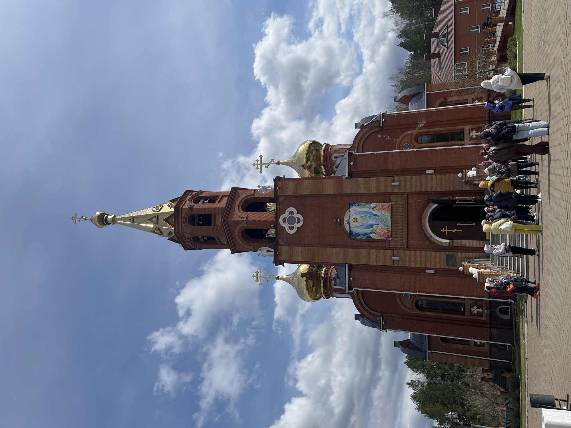 The image captures an Eastern Orthodox church in Chaikovsky, Russia, on a bright, partially cloudy day. The prominent subject is a grand red-brick church featuring multiple golden onion domes and a tall, elegant spire topped with a golden Orthodox cross. Additional golden crosses adorn other domes. The church facade displays intricate architectural details, including arched windows and doorways, a quatrefoil decorative element, and a large, colorful mosaic or fresco above the main entrance depicting religious figures, likely the Virgin Mary and saints.
In front of the church, a group of people, mostly adults, are gathered on a paved pathway made of interlocking pavers. They appear to be standing in a line or queue, possibly waiting to enter the church or attending an outdoor event. Their attire suggests casual outerwear appropriate for moderate weather.
In the background, behind the church, another red-brick building with multiple windows is visible, along with some trees. The sky is bright blue with scattered white, fluffy clouds, indicating fair weather during the daytime, likely midday or afternoon given the clear light. No discernible text is visible on the church or in the scene. The overall atmosphere is calm and orderly.