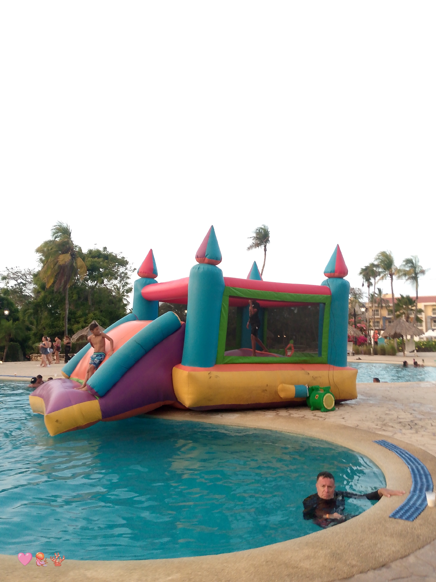 The image shows an inflatable bounce house and slide partially in a swimming pool. A boy is positioned on the slide about to enter the water. Another child is inside the bounce house. A man is also in the pool near the edge. The scene appears to be in a resort or hotel area with a pool. Palm trees and other vegetation are visible in the background, along with what appears to be resort buildings. People can be seen in the background. The location is Playa El Agua, Venezuela.