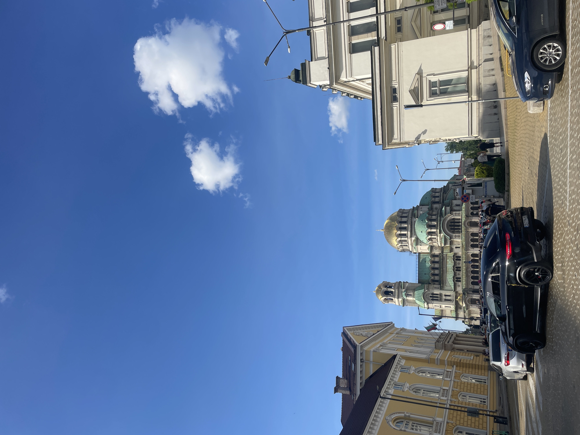 The image shows a street scene in Sofia, Bulgaria. In the background is the Alexander Nevsky Cathedral, recognizable by its golden dome. The photo is taken from street level, with the cathedral and the tops of surrounding buildings occupying the lower portion of the frame, and a mostly clear blue sky with a few scattered clouds filling the upper portion. Several cars are parked along the street, including black and white ones. The buildings are diverse in style, with light-colored facades and architectural details. A few street lights are visible, and a few people are discernible on the street, though the distance makes it hard to discern details about them.
