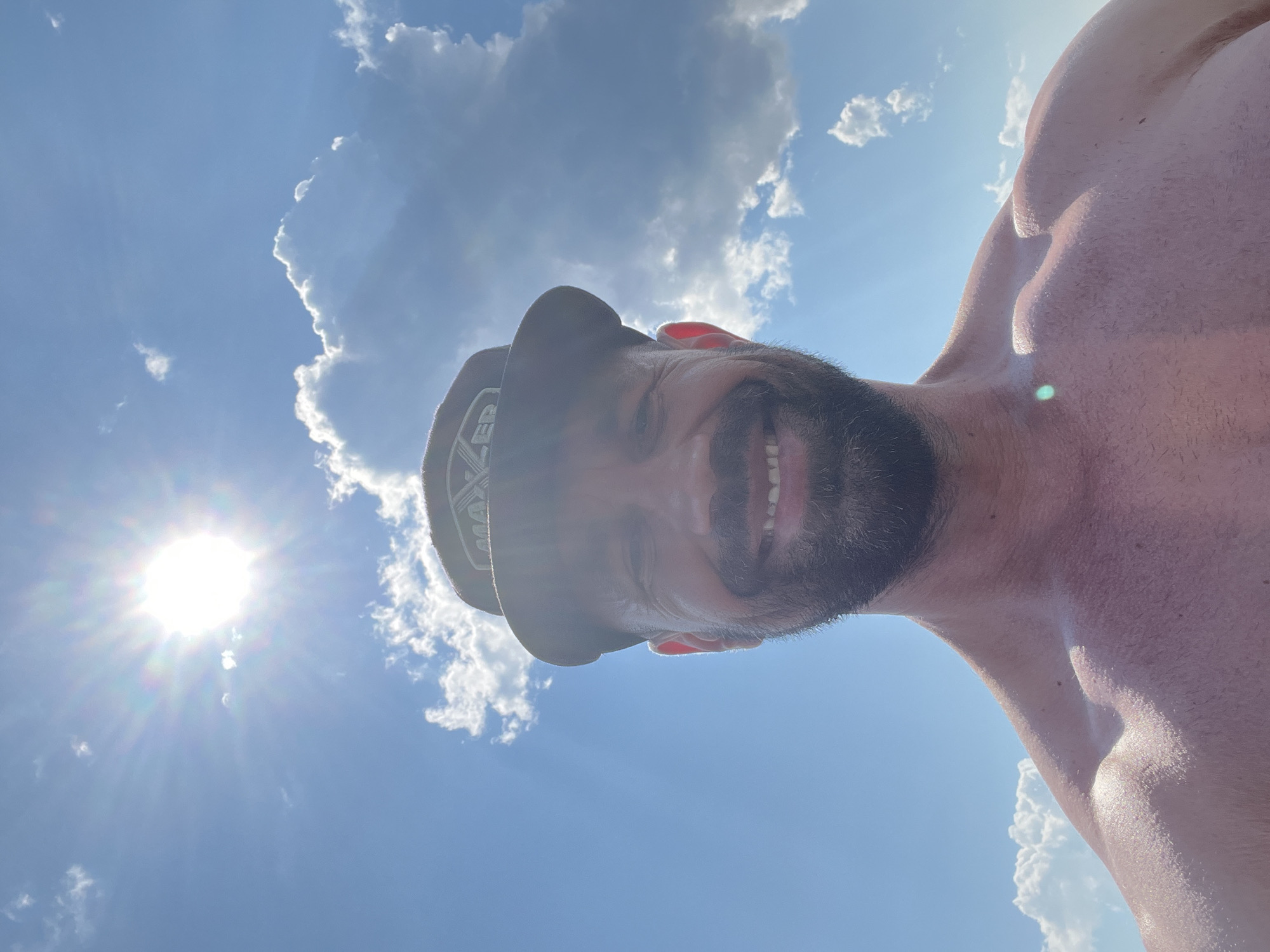 A man with a beard and a hat smiles at the camera. He is shirtless, with sun glinting off his shoulders and chest. He appears to be outside, with a bright sun and blue sky with white clouds in the background. The location is the city of Sosnovka, Russia.
