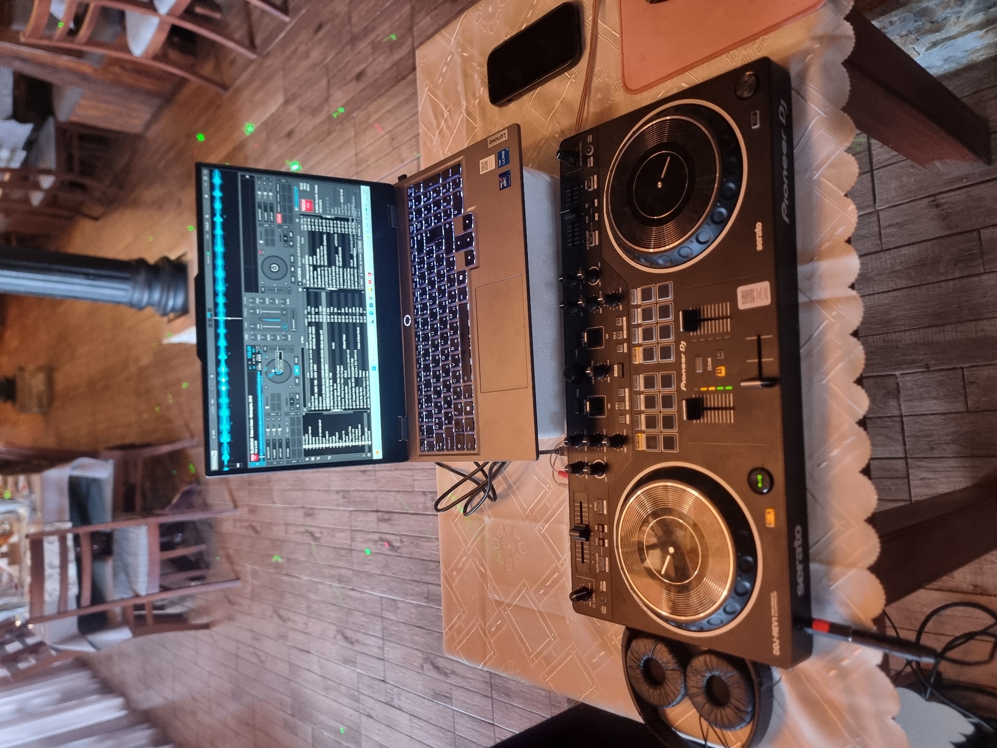 A DJ setup, comprising a Lenovo laptop displaying DJ software, a Pioneer DDJ-REV1 controller, and headphones, is placed on a table covered with a patterned white tablecloth. A smartphone and a pink object are also on the table. The setting appears to be indoors in Banichan, Bulgaria, considering the location indicated. The room's interior includes wooden furniture and decor elements. There are no people visible in the frame. The focus is on the DJ equipment and its arrangement.