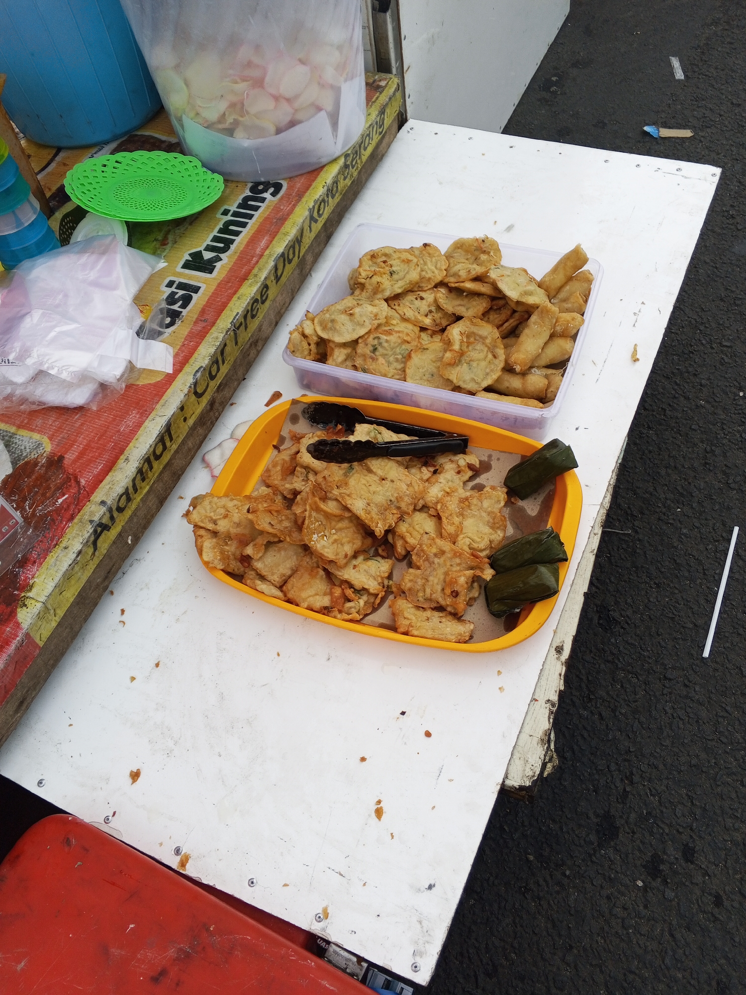 The image shows a street food stall in Serang, Indonesia. The stall features a white table laden with food items. In a clear plastic container, there are fried tempeh chips and other fried snacks. Next to it, a yellow tray holds more fried tempeh and three wrapped food items that appear to be vegetables encased in banana leaves. Black tongs are placed on the yellow tray. In the background, a blue bucket and a clear plastic bag filled with what looks like dried flower petals are visible. A wooden plank with yellow and red writing, partially obscured, runs along the edge of the table, with text including "Alamari: Car Free Day Kota Serang". The table is positioned on asphalt.