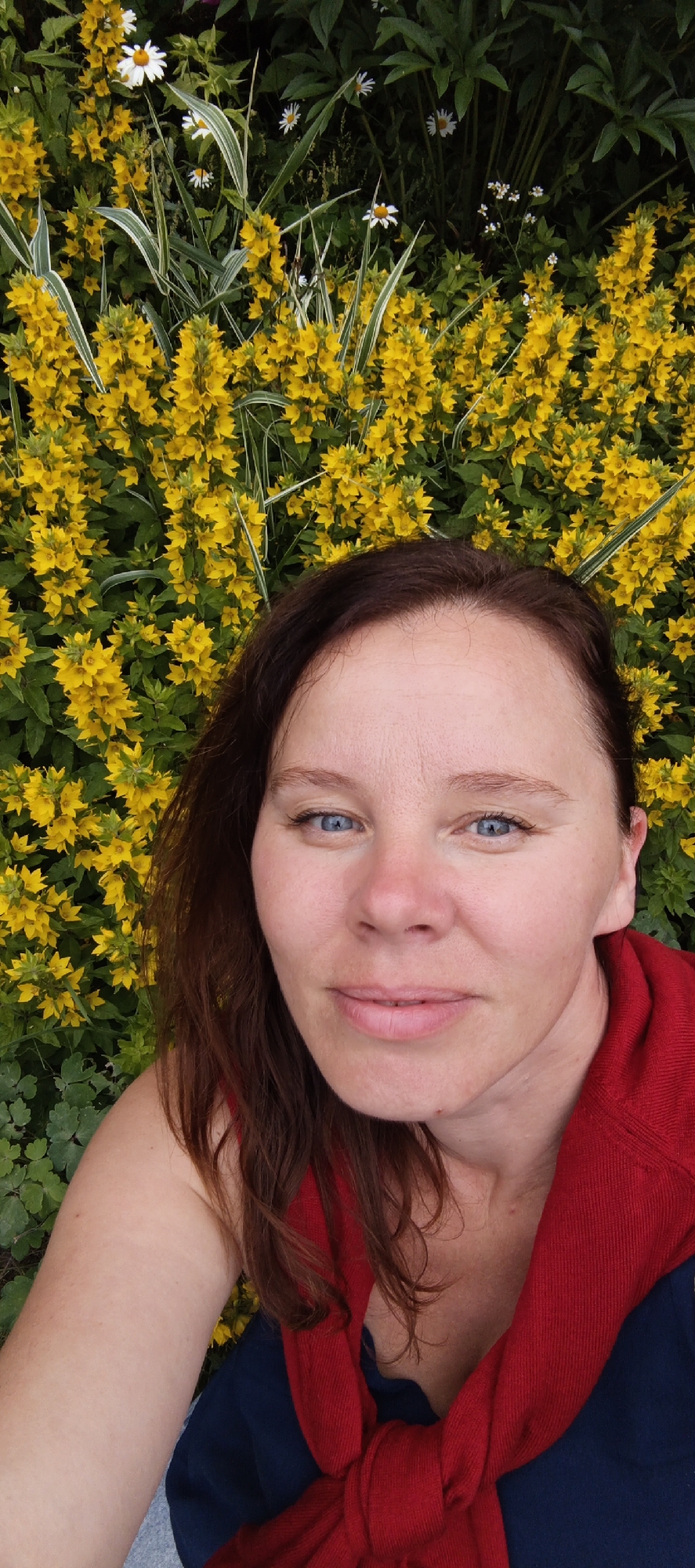 A woman with auburn hair and blue eyes is taking a selfie outdoors, with a backdrop of bright yellow flowers and green foliage. She is wearing a red sweater tied around her neck and shoulders over a dark blue top. The image appears to be taken in a garden or park setting. The exact location in Chaikovsky, Russia, is not discernible from the image itself.