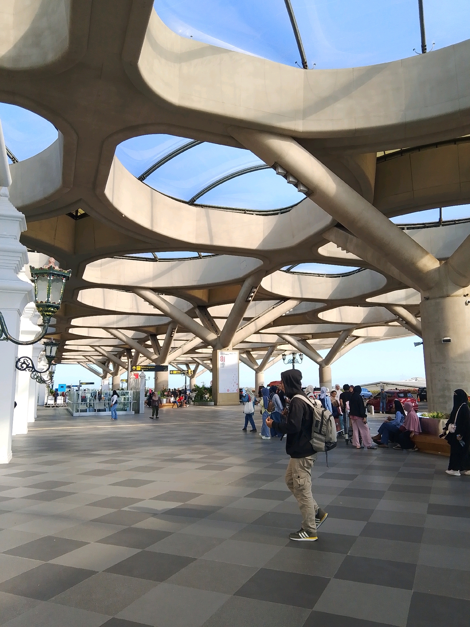 The image depicts an outdoor area with a distinctive architectural design featuring a large, multi-layered concrete canopy with an organic, wave-like structure supported by numerous branching columns. The canopy has large circular openings that allow sunlight to filter through, illuminating the checkered tiled ground below. In the foreground, a person wearing a black hooded jacket, khaki cargo pants, and athletic shoes walks from left to right, carrying a backpack. Other people are visible in the background, some standing, some sitting on a bench, and others walking. There are also signs indicating directions and what appears to be an airport terminal in the distance, suggesting the location is an airport in Palihan, Indonesia. The overall environment is bright with a clear blue sky visible through the canopy's openings.