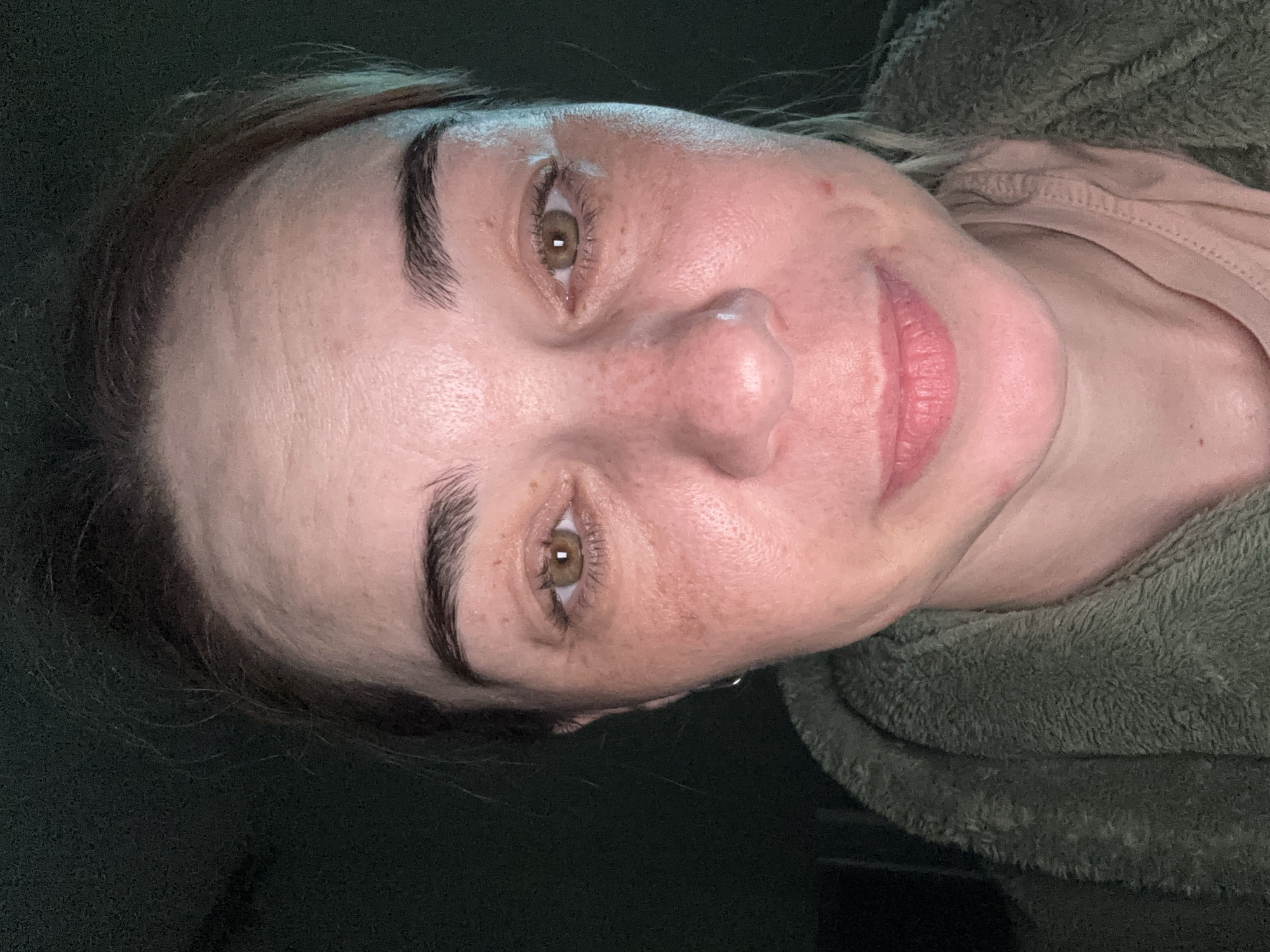 The image shows a close-up portrait of a woman with fair skin and hazel eyes. She has dark eyebrows and her hair appears to be pulled back. Her face shows some freckles and minor skin imperfections. She is wearing a light pink shirt or top and is partially covered by what appears to be a green, fuzzy jacket or blanket. The background is dark and out of focus. The location and date are unknown.