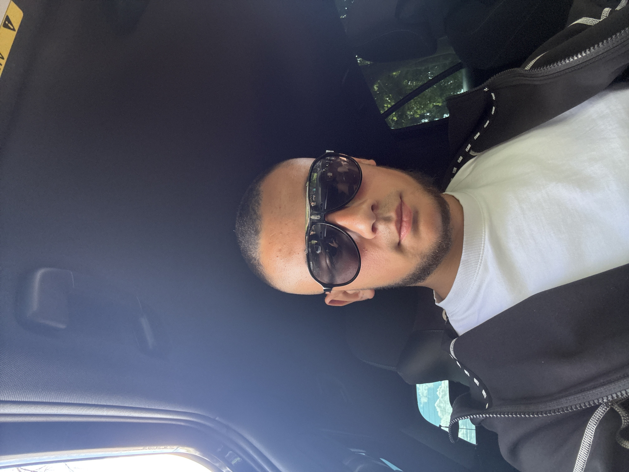 A man wearing sunglasses, a white t-shirt, and a black jacket is in a car with a black interior. The location in the city of Elhovo, Bulgaria is unknown. The image is taken from an overhead perspective inside the car.